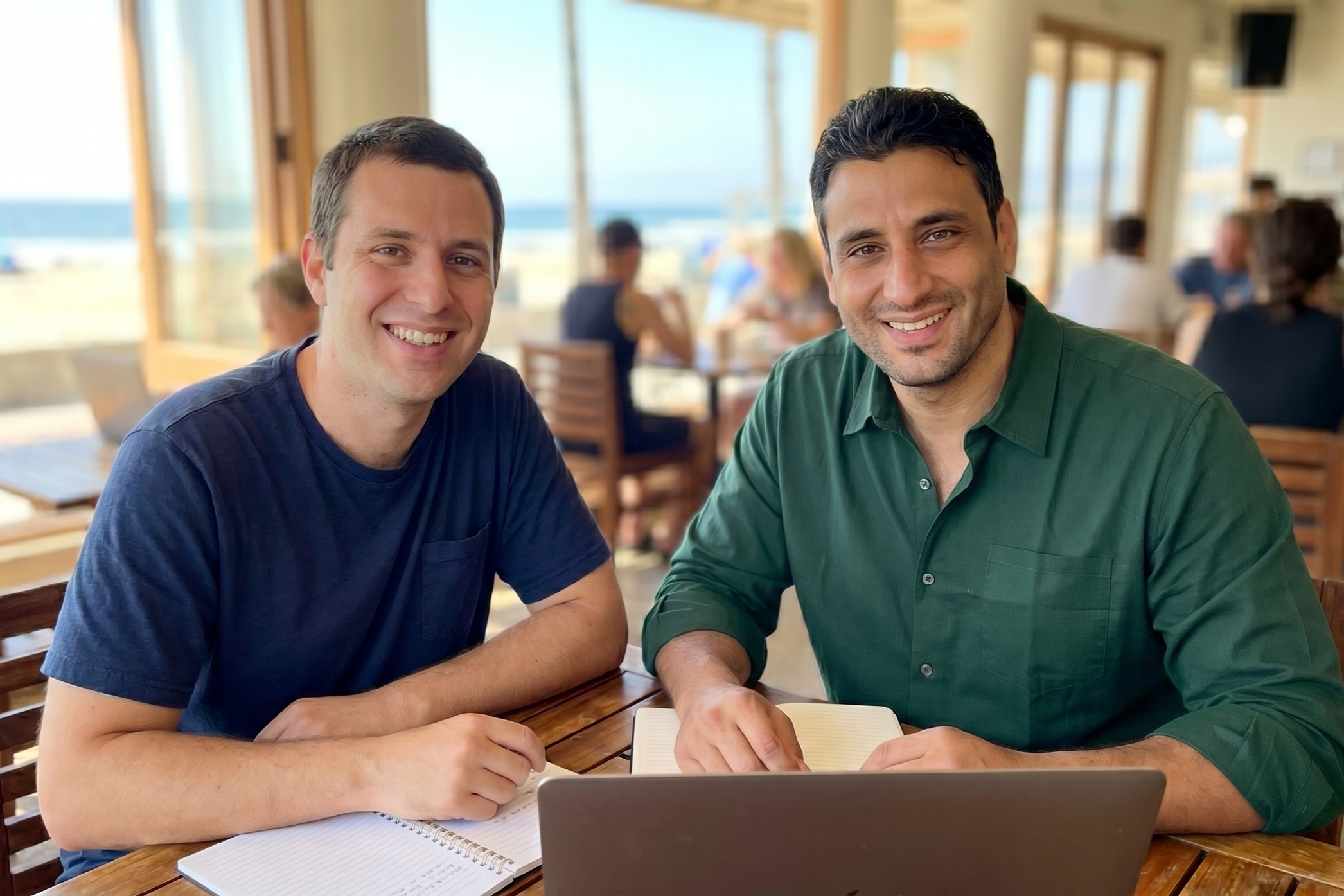 Two smiling men sitting at a wooden table in a bright cafe with large windows, working on laptops and taking notes.