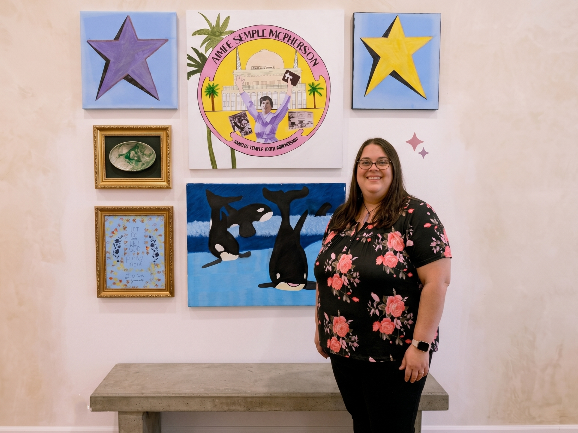 A woman with glasses and long brown hair, smiling, standing in front of a decorated wall with various colorful art pieces. The wall includes paintings of stars, a circular graphic with a woman holding a book, a whale, and a framed quote reading 'Let go & let God pray more love'. The woman is wearing a black shirt with pink floral patterns, and a smartwatch on her left wrist.