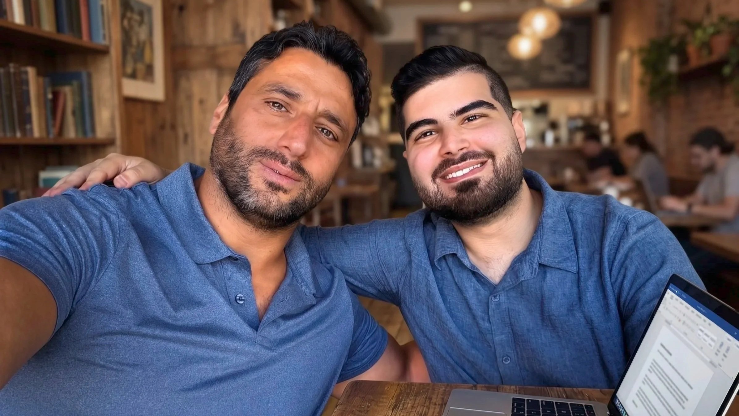 Two men taking a selfie in a cozy, wood-paneled cafe, smiling, with a laptop in front of them and books on shelves in the background.