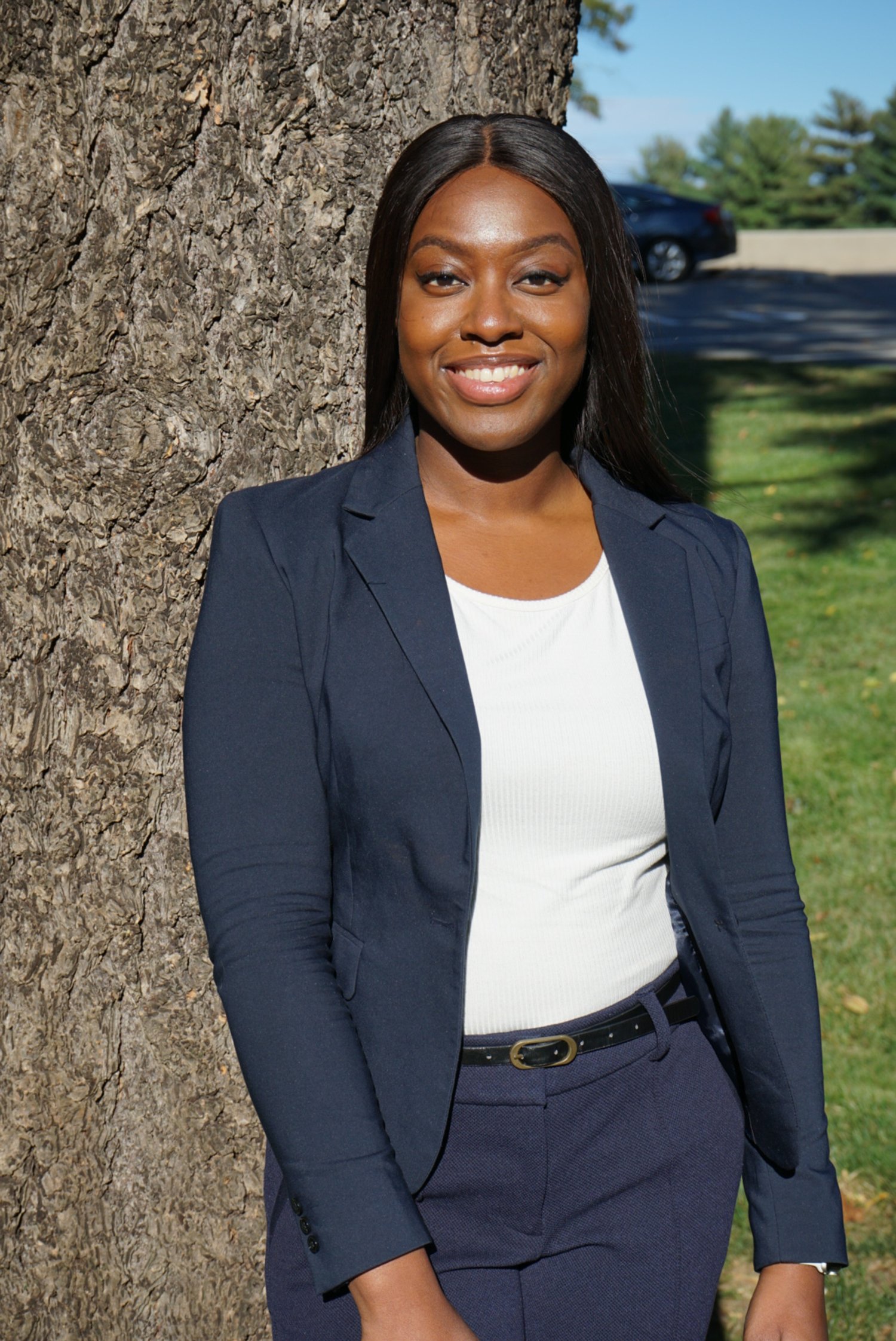 Young woman with glowing, dark skin, brown eyes, and long black hair is standing in front of a tree and looking into camera. She is wearing a navy pantsuit and white blouse.