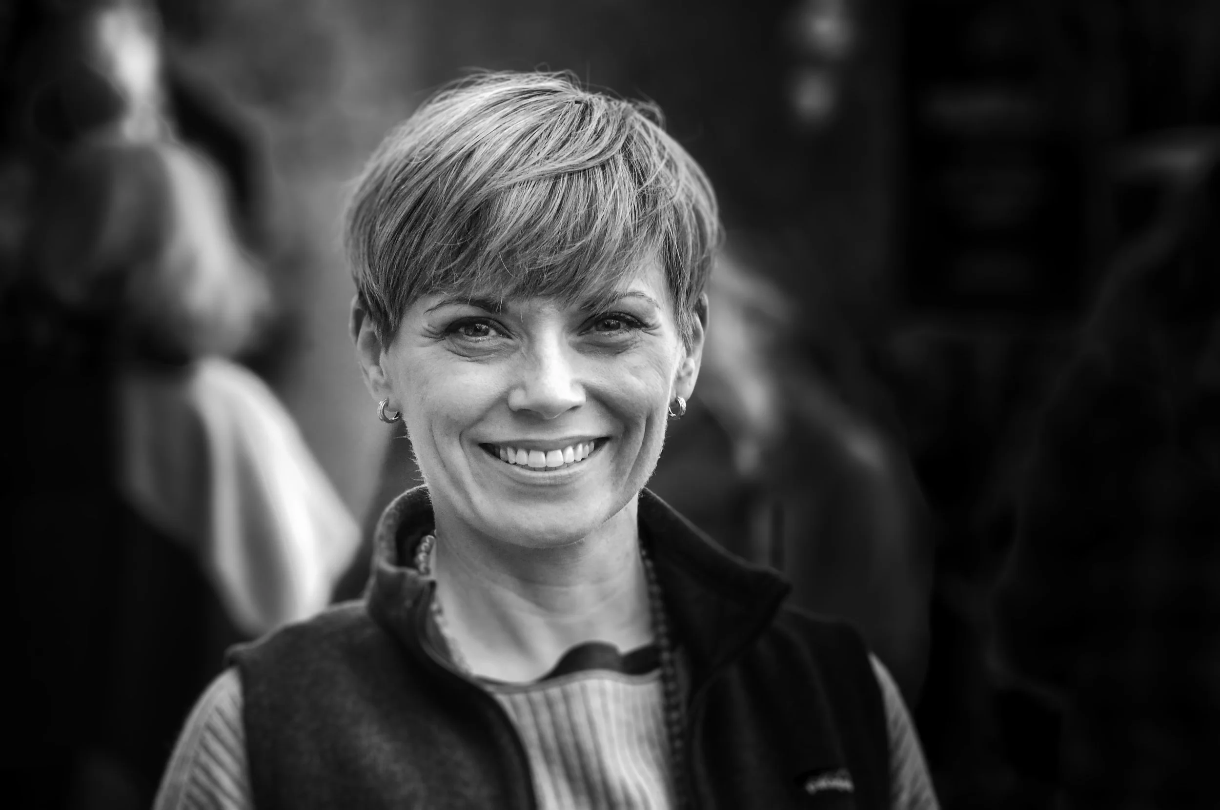 Black and white image of woman standing outdoors. She has pale skin and short hair. She is smiling and facing the camera.