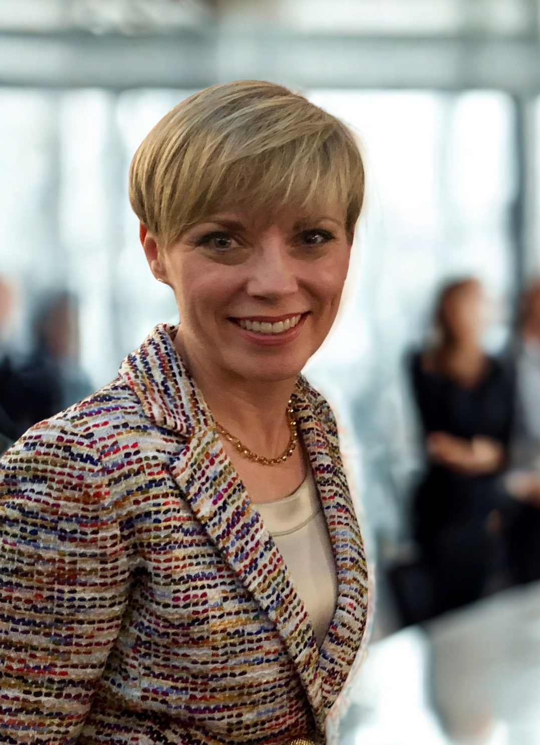 A smiling woman with short blonde hair wearing a striped blazer and jewelry, standing in a well-lit room with blurred people in the background.