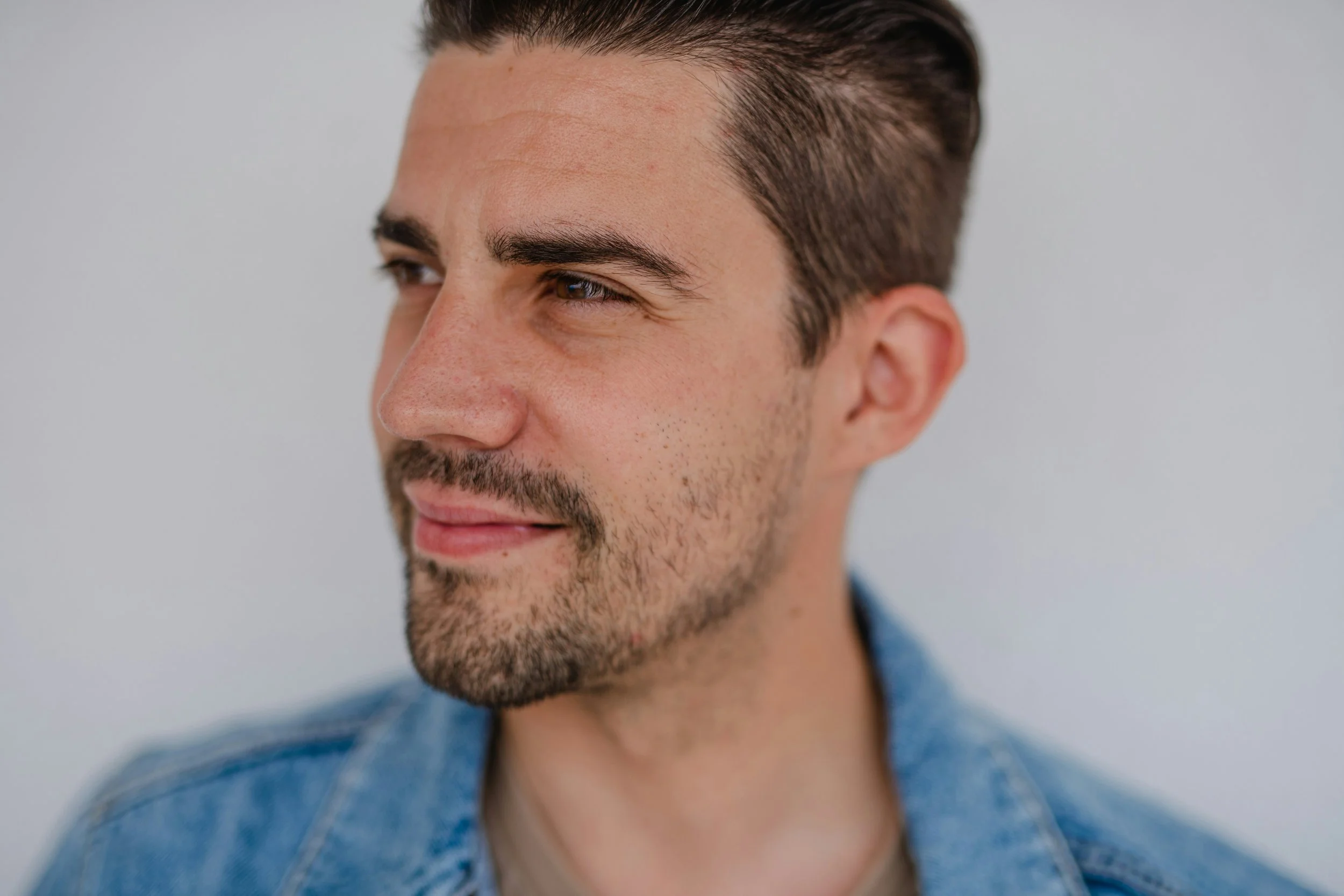 Close-up of a man with short brown hair, light skin, a beard, and mustache, wearing a denim jacket, looking to his left with a slight smile, against a plain light background.