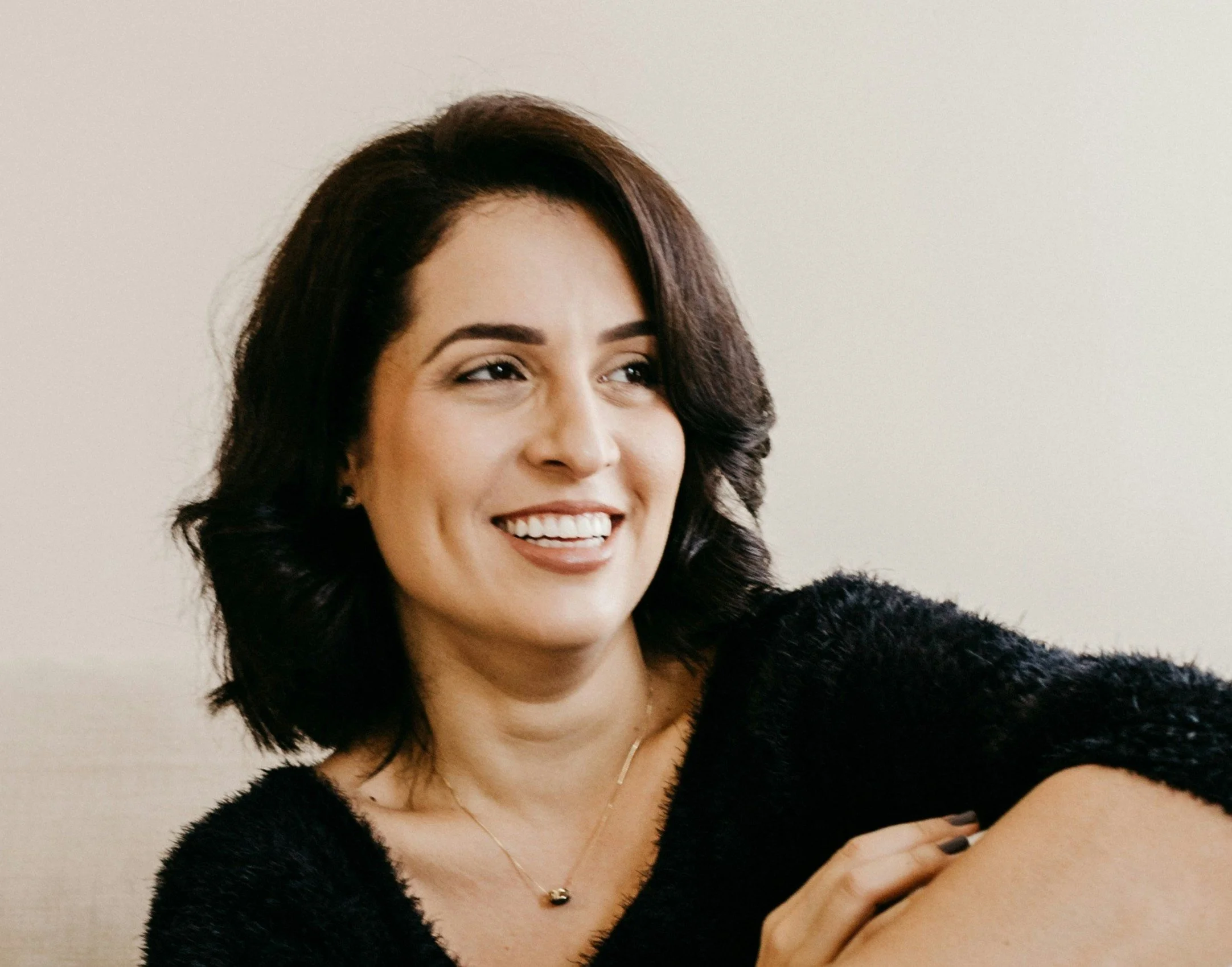 A woman with dark brown hair smiling, wearing a black fuzzy sweater and a delicate gold necklace, sitting against a light background.