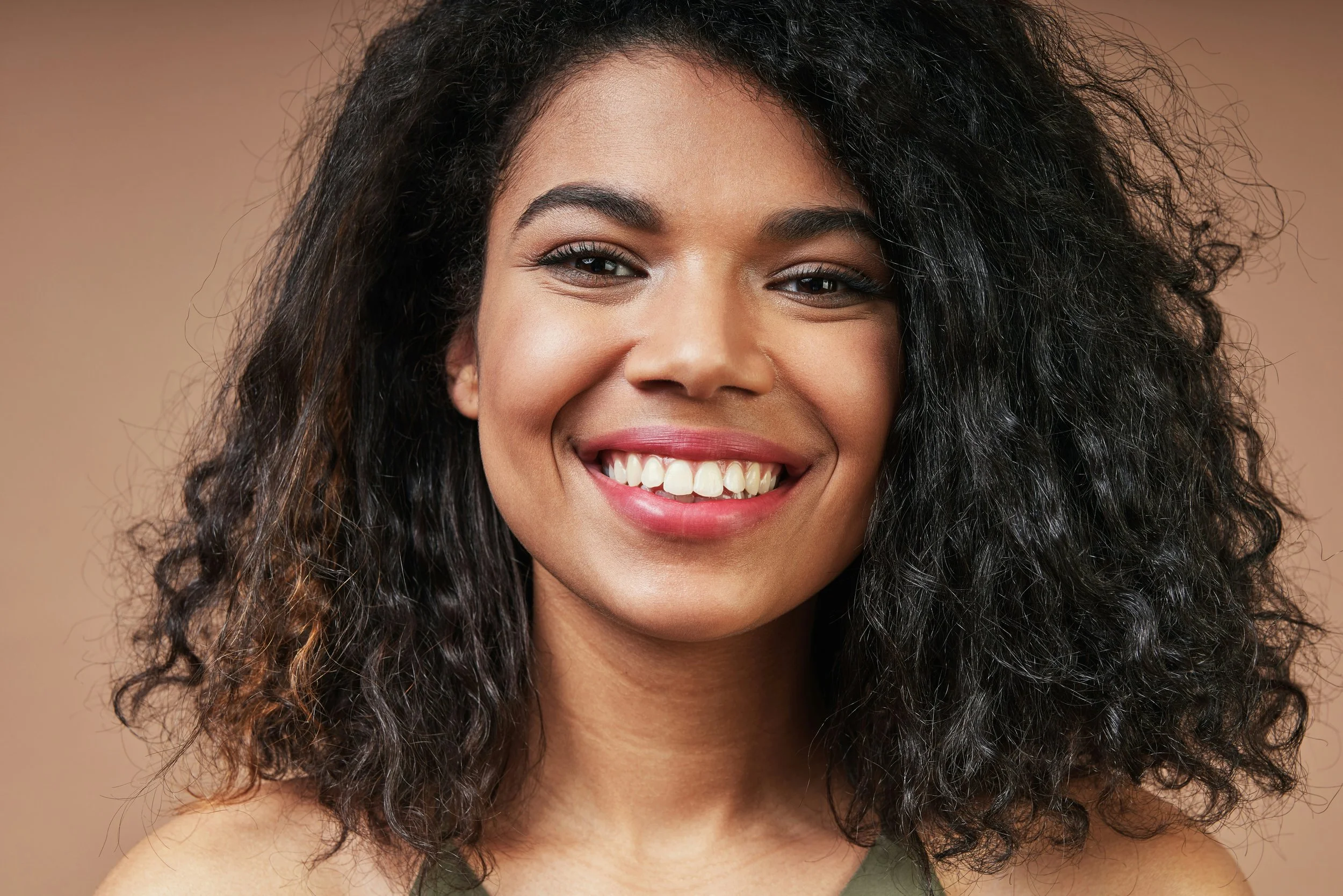 Close-up of a smiling woman with curly black hair, light brown skin, makeup, and a green top against a neutral background.