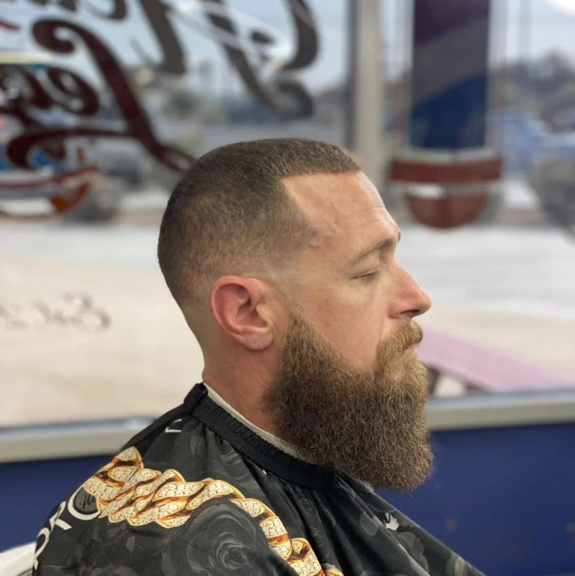 Profile of a man with short hair and a full beard sitting with his eyes closed in a barbershop.