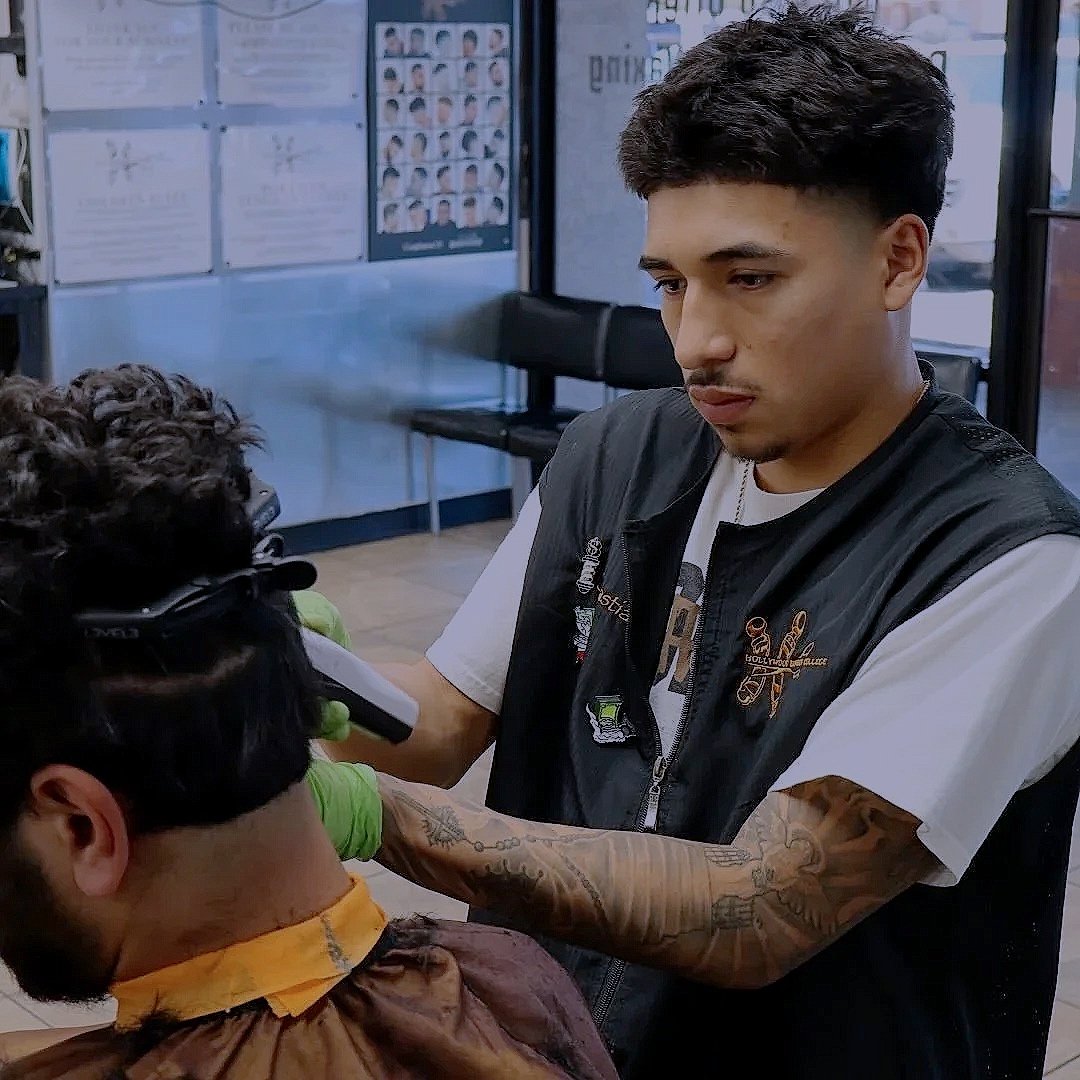 A young hairstylist with tattoos on his arms and wearing gloves is giving a haircut to a man with curly hair inside a barbershop or salon.