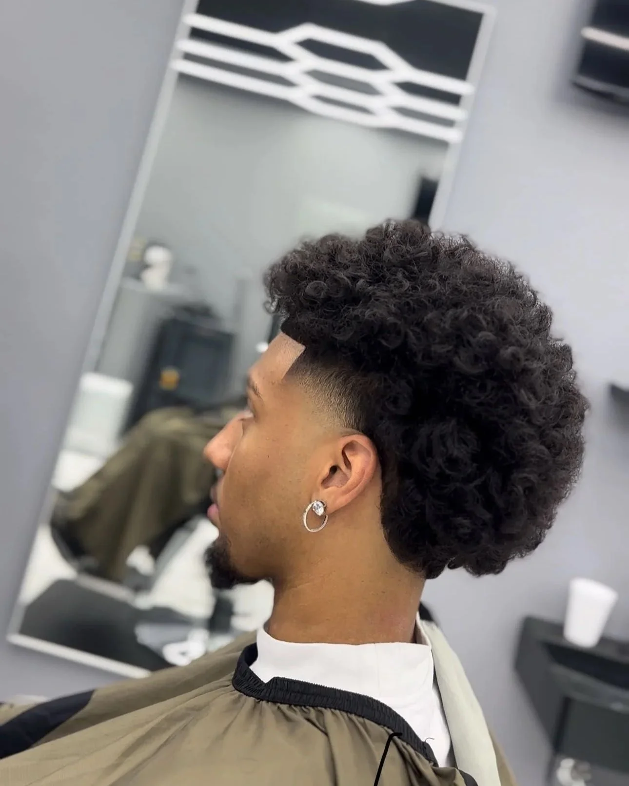 Side view of a man at a barber shop with a fresh curly hairstyle and a fade, wearing an earring and a barber smock, in front of a mirror.