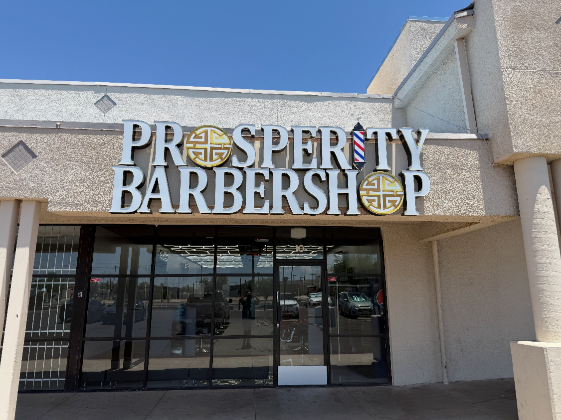 Exterior of a barbershop named Prosperity Barbershop with a sign featuring Chinese filter and barber pole symbols, located in a shopping plaza on a sunny day.