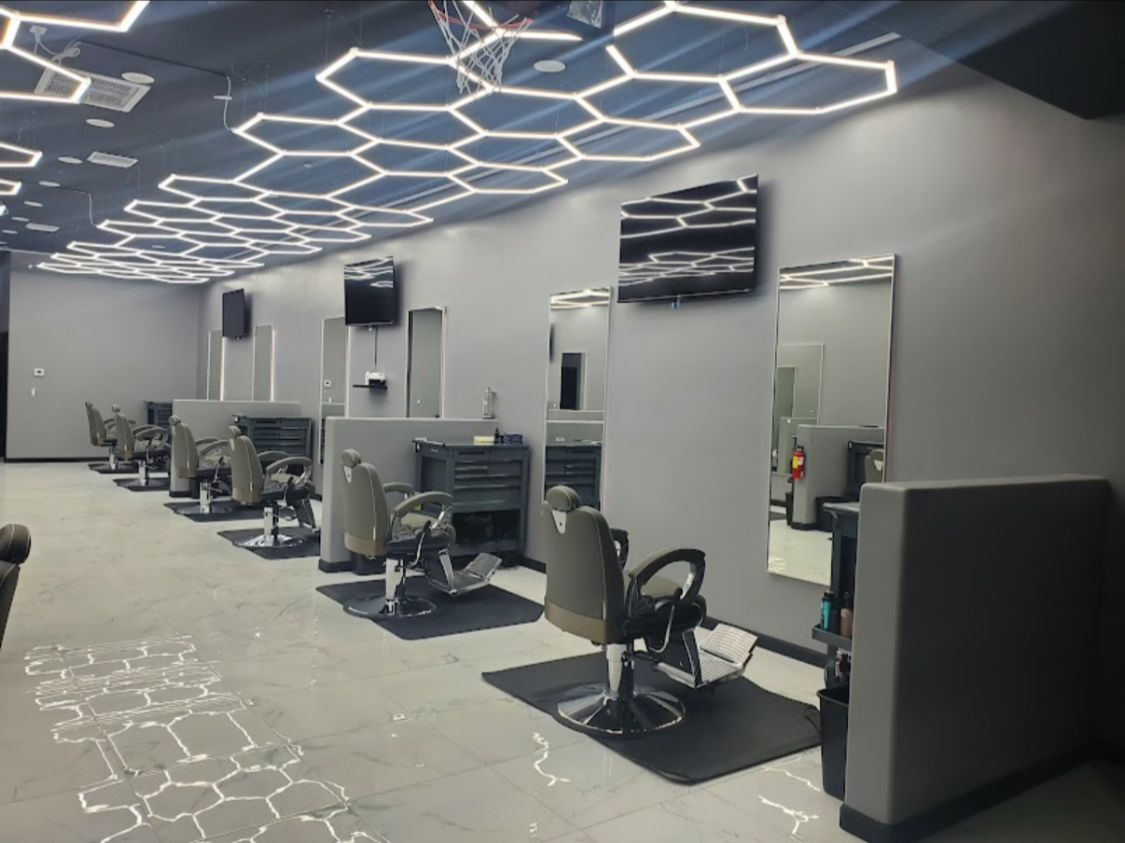 A modern hair salon with several empty styling chairs, mirrors, and TV screens mounted on the wall. The ceiling features a geometric LED light design, and the floor has white tiles with hexagon-shaped patterns.