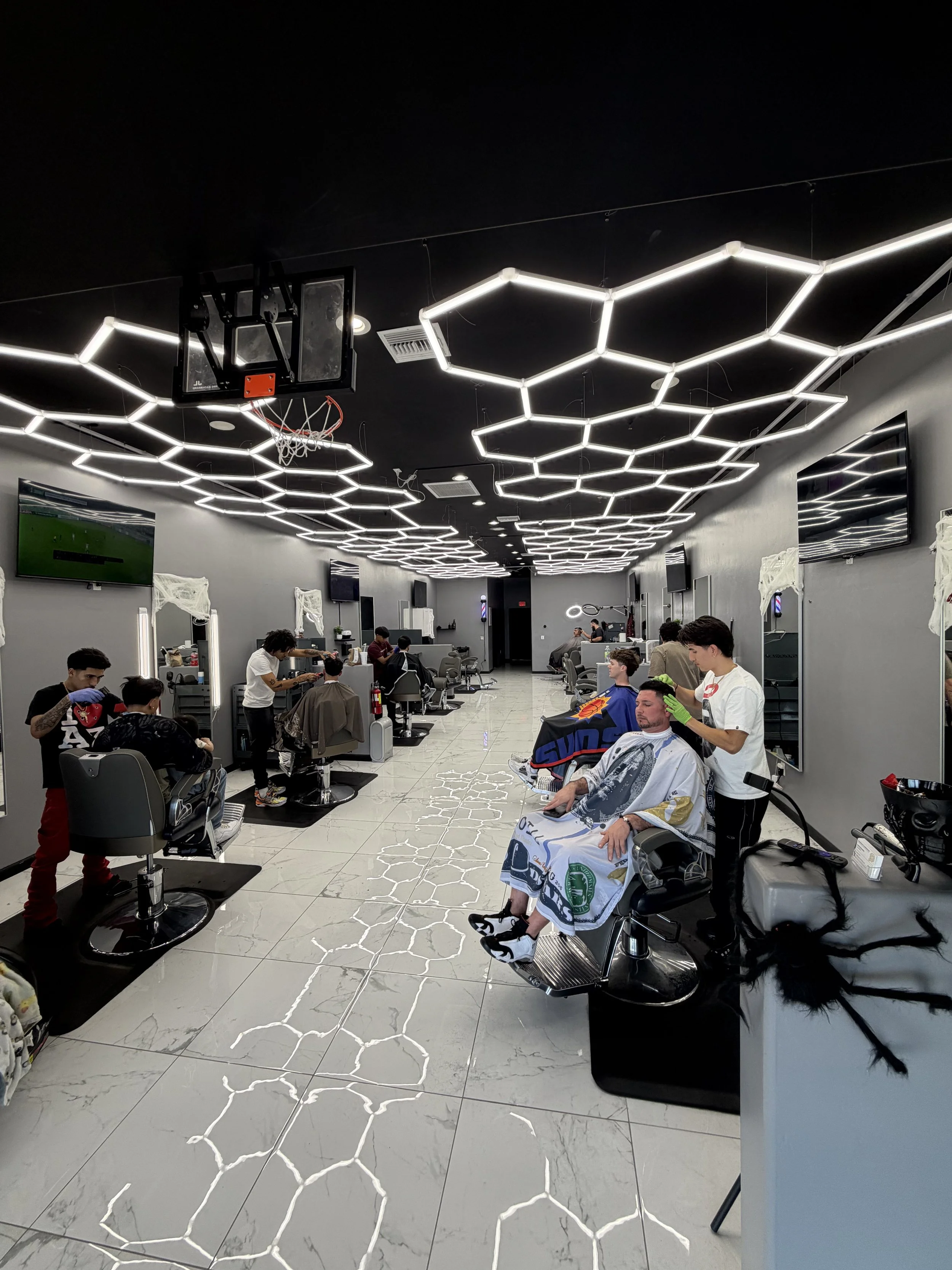 Modern barbershop with multiple customers getting haircuts, LED hexagon ceiling lights, and sleek gray and white decor.