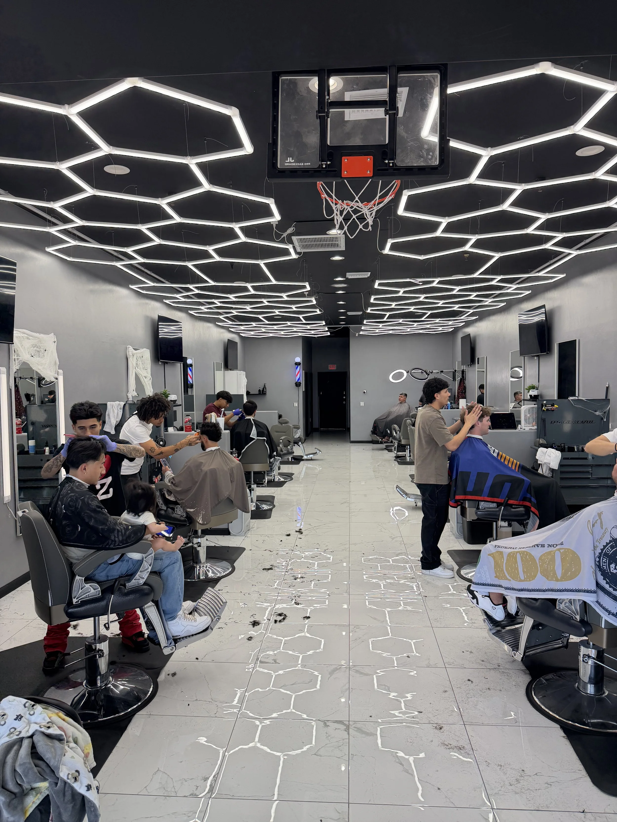 Modern barbershop with multiple clients getting haircuts, sleek black ceiling with hexagonal light fixtures, and a basketball hoop mounted on the ceiling.