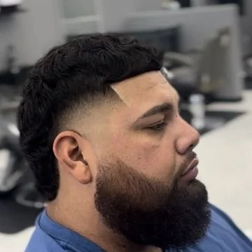 Side profile of a man with a fade haircut and full beard, seated in an indoor setting.
