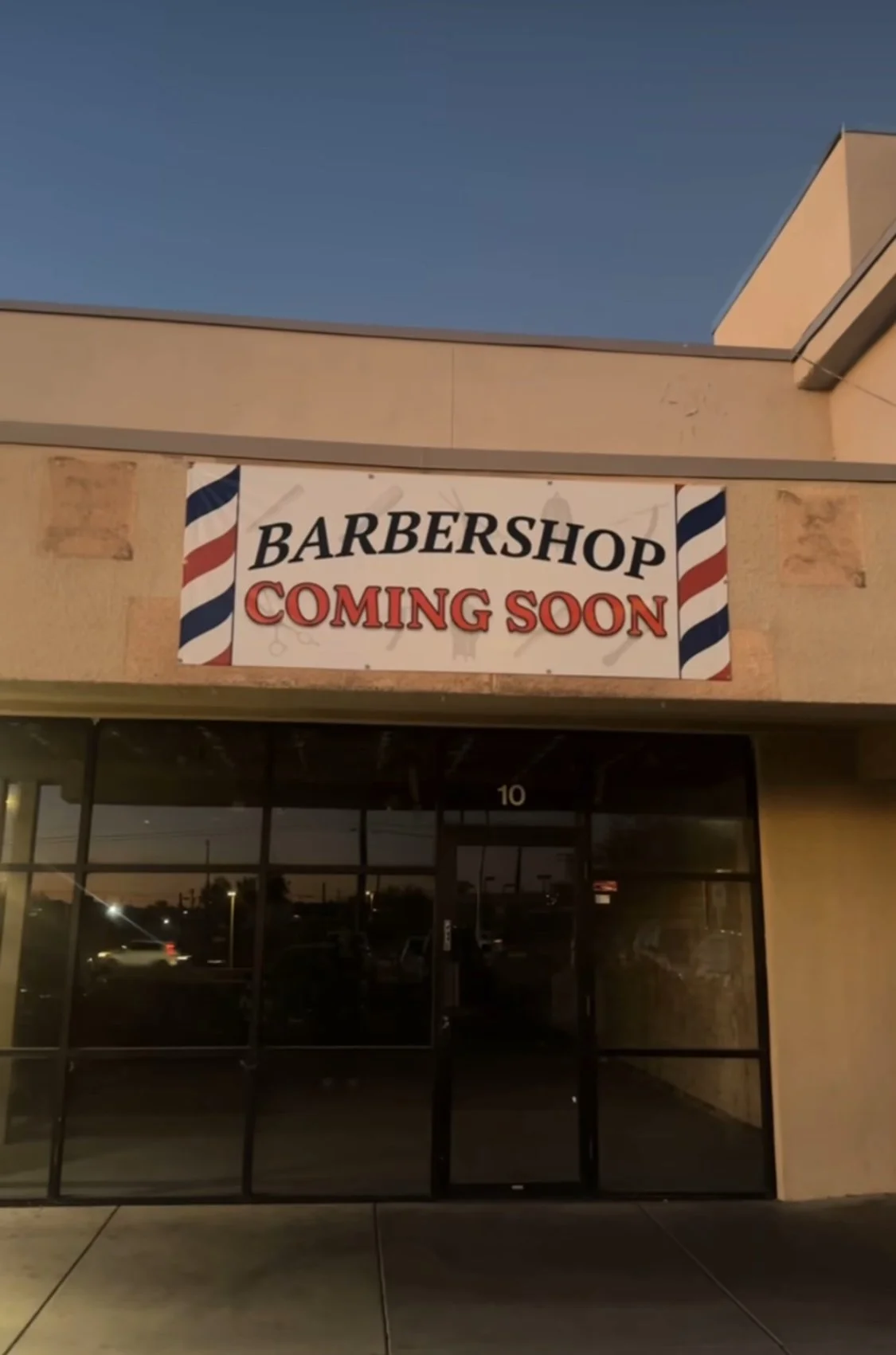 A storefront with a sign that reads 'Barbershop Coming Soon' and features barber pole graphics on either side, with the storefront door and windows below.