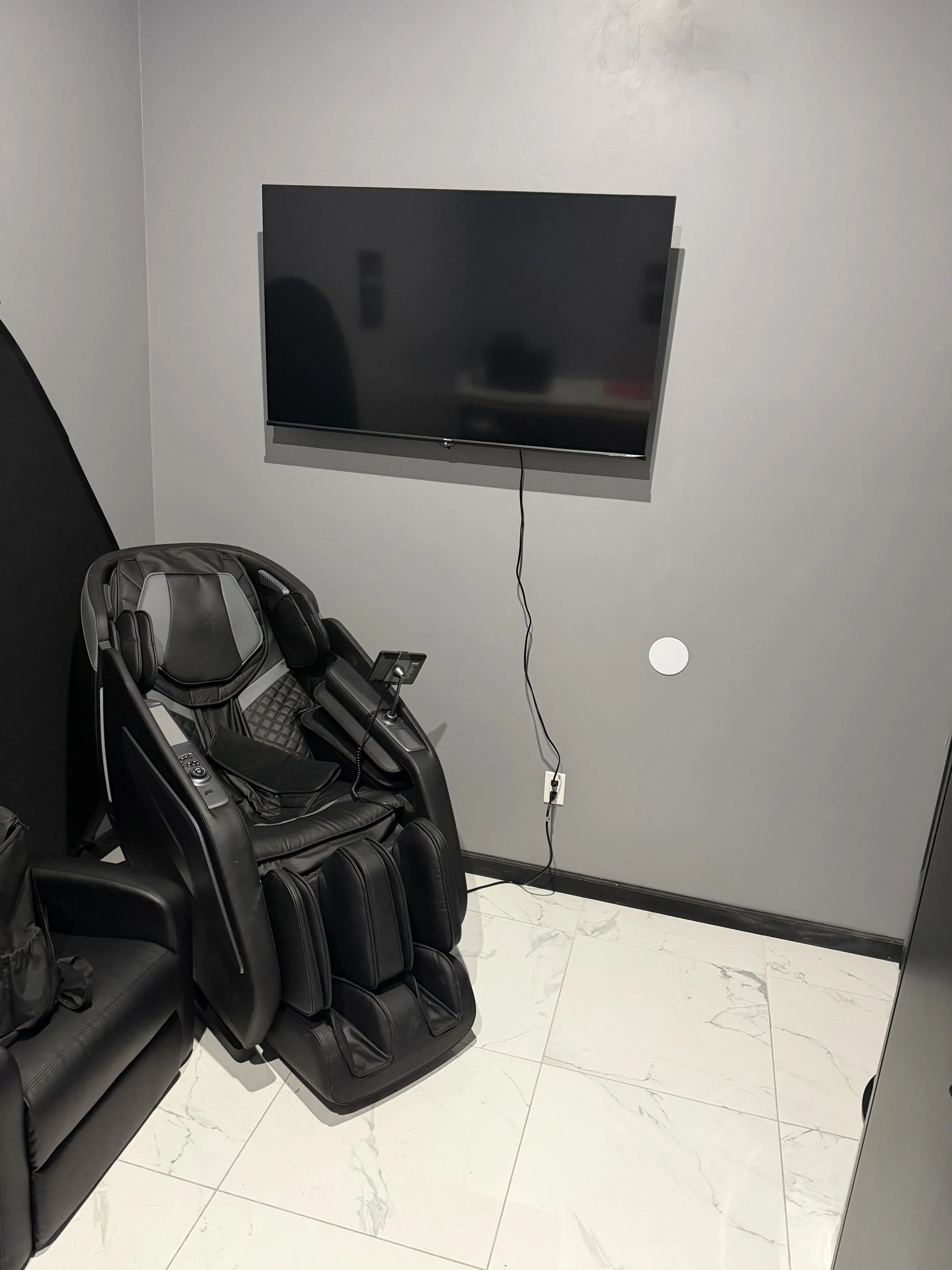 A room with a mounted flat-screen TV on the wall, a massage chair with electronic controls, and a black leather couch, all on a white marble floor.