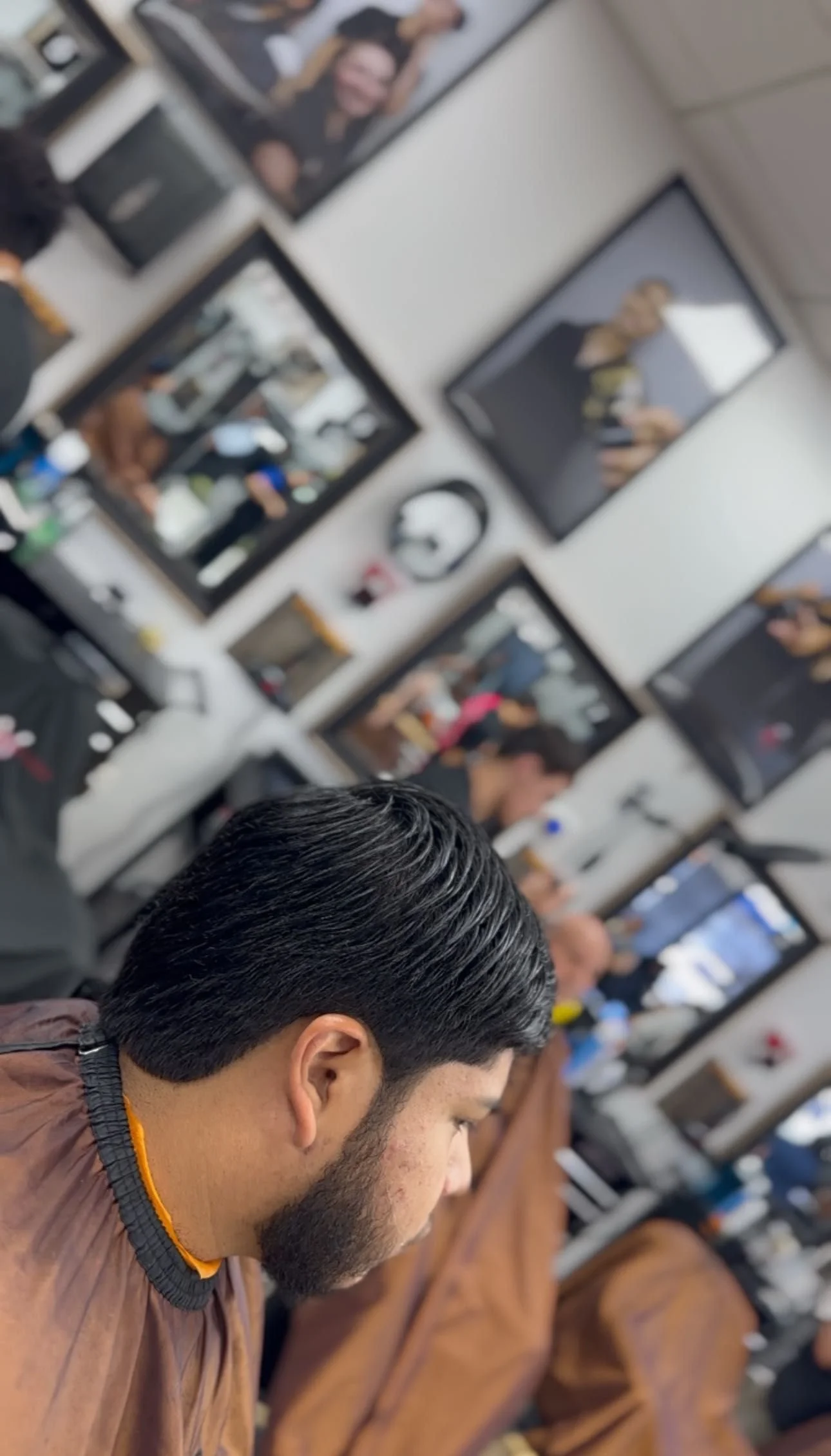 A man with black, slicked-back hair and a beard is sitting in a barbershop, looking down. Multiple mirrors and other patrons are visible in the background, with some people receiving haircuts.