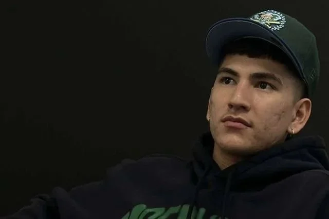 A young man wearing a dark hoodie and a black baseball cap with a logo, sitting against a black background.