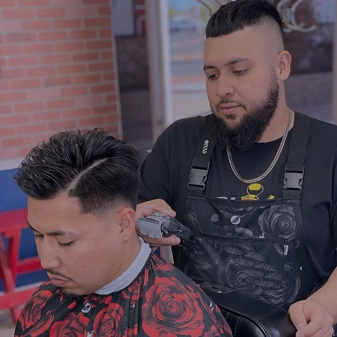Barber giving a man a haircut with clippers in a salon with a brick wall and large windows in the background.