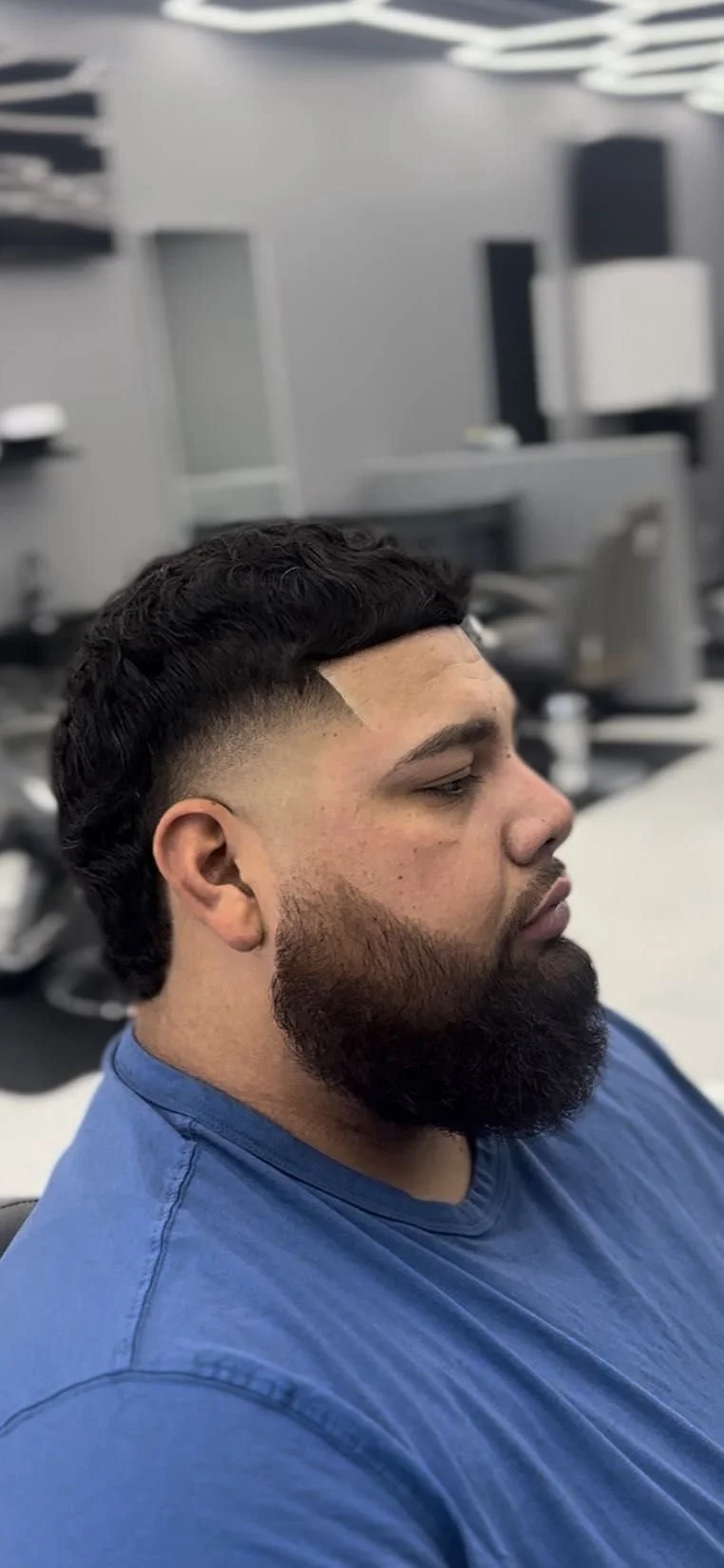 Side view of a man with a tapered haircut and full beard, wearing a blue shirt, in a hair salon or barbershop.