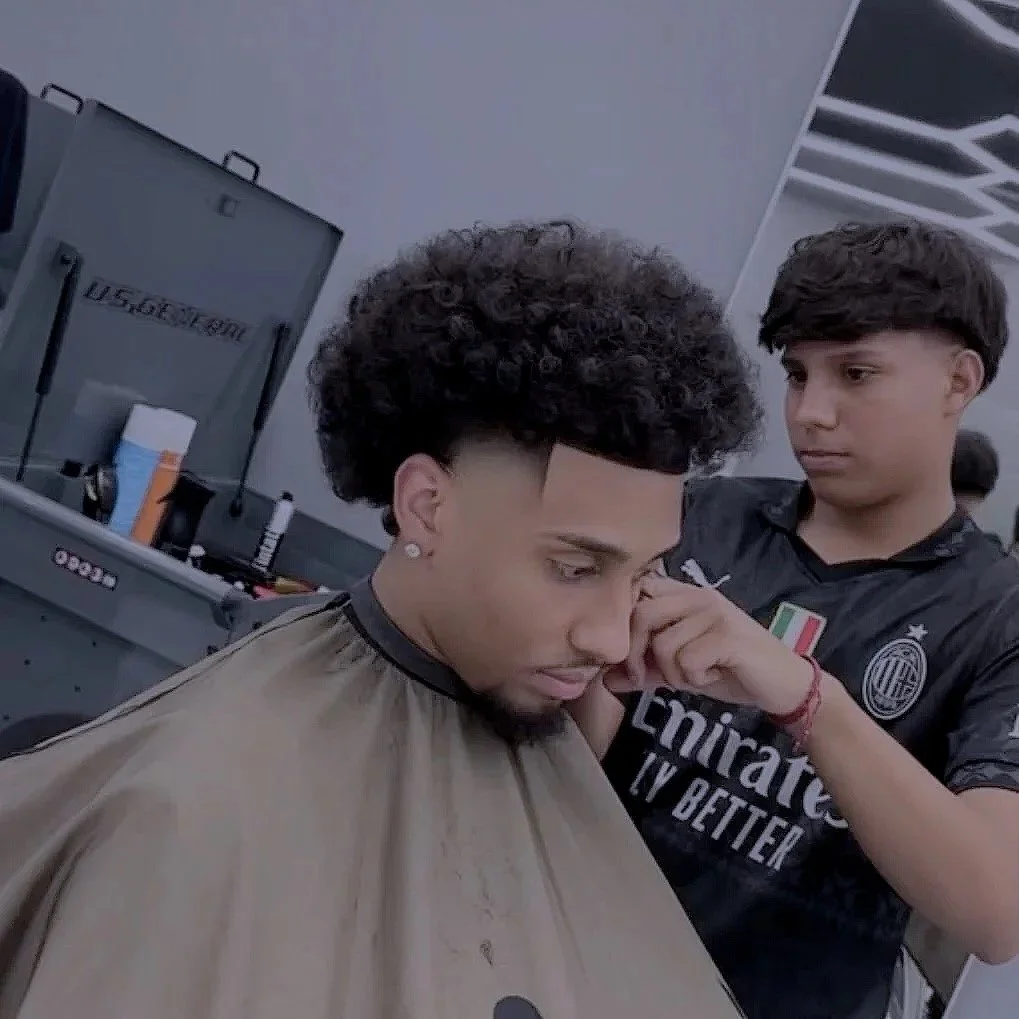 A young man getting a haircut at a modern barber shop, with a barber standing nearby in a black Milan football jersey.