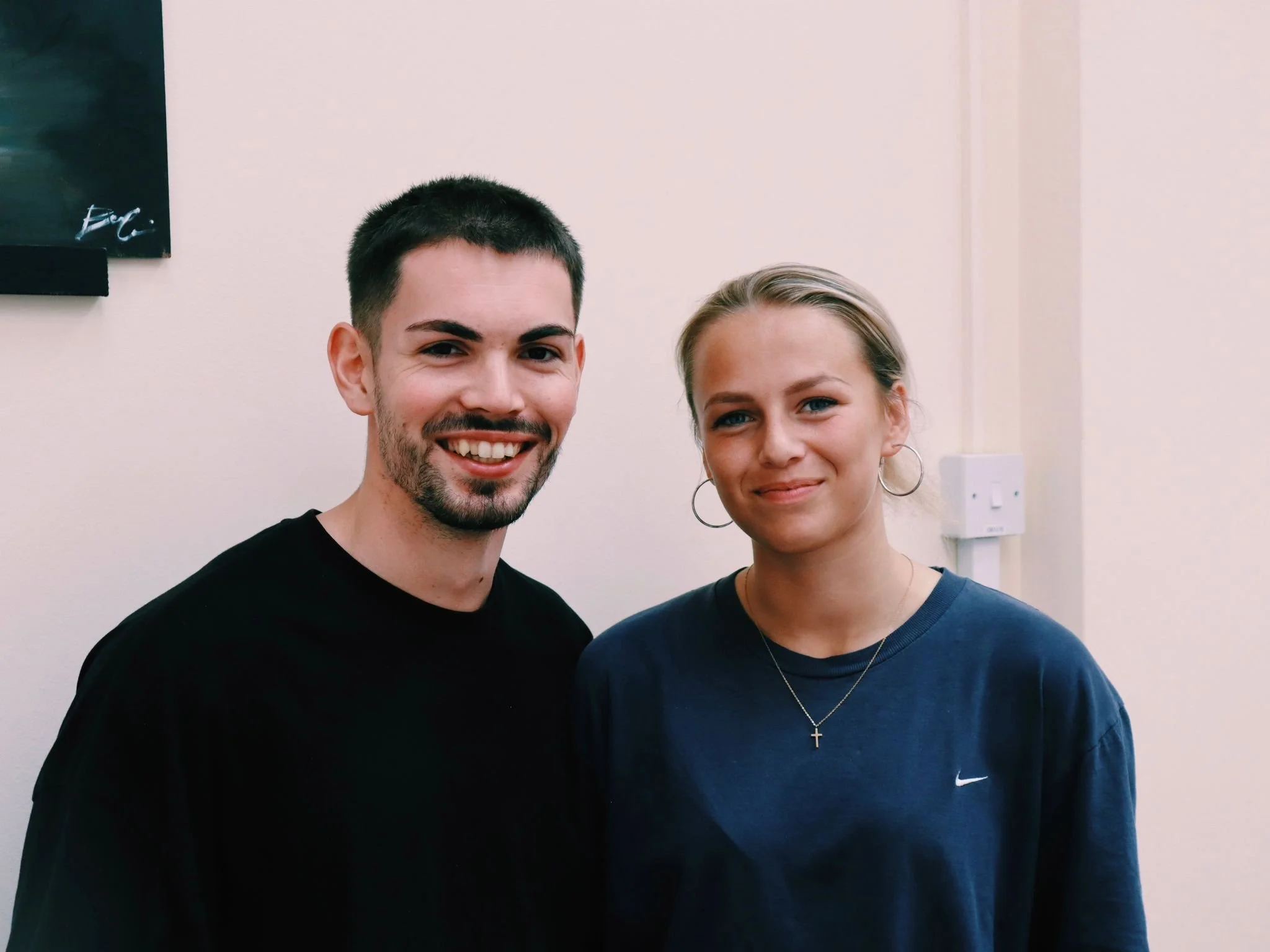 A young man with short dark hair and a beard smiling, standing next to a young woman with blonde hair tied back, wearing hoop earrings and a cross necklace, both standing indoors against a beige wall.