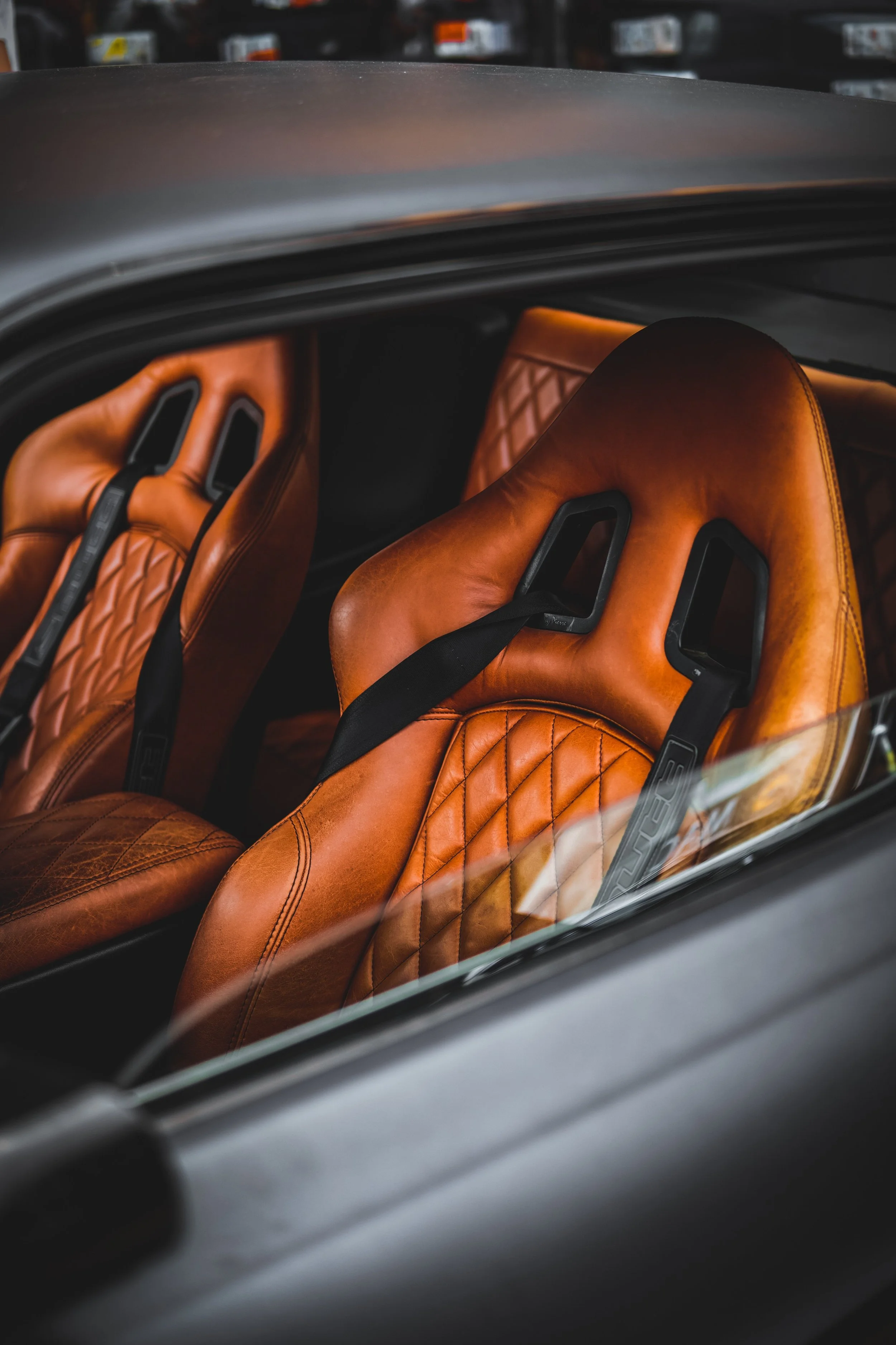 Close-up of two brown leather racing car seats with black harnesses inside a sports car.