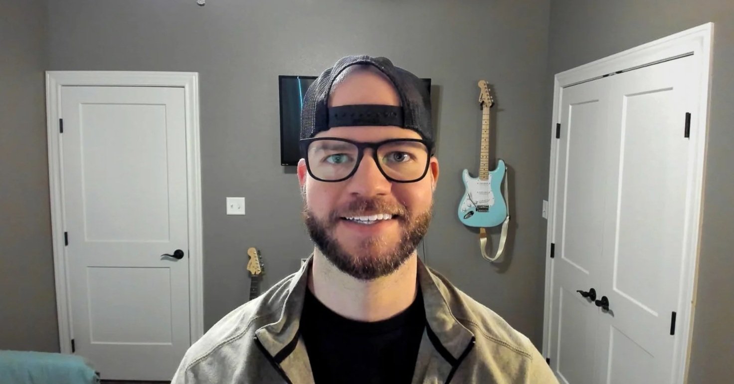 A man with a beard and glasses wearing a black cap backwards and a beige jacket, smiling in a room with gray walls and two guitars hanging on the wall behind him.