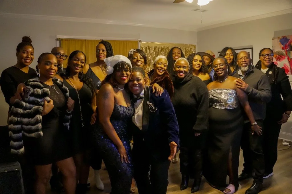 Group of women and men in formal and semi-formal attire posing together indoors during a celebration.