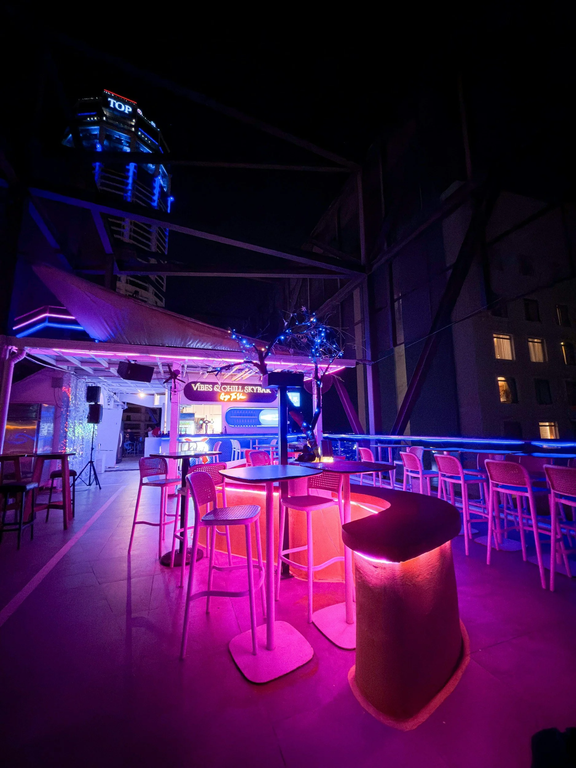Night view of a vibrant rooftop bar with neon pink and blue lighting, bar stools, and a sign reading 'Vibes & Chill Skybar'. In the background, a building with lit windows and a tower with the word 'TOP' illuminated at the top.