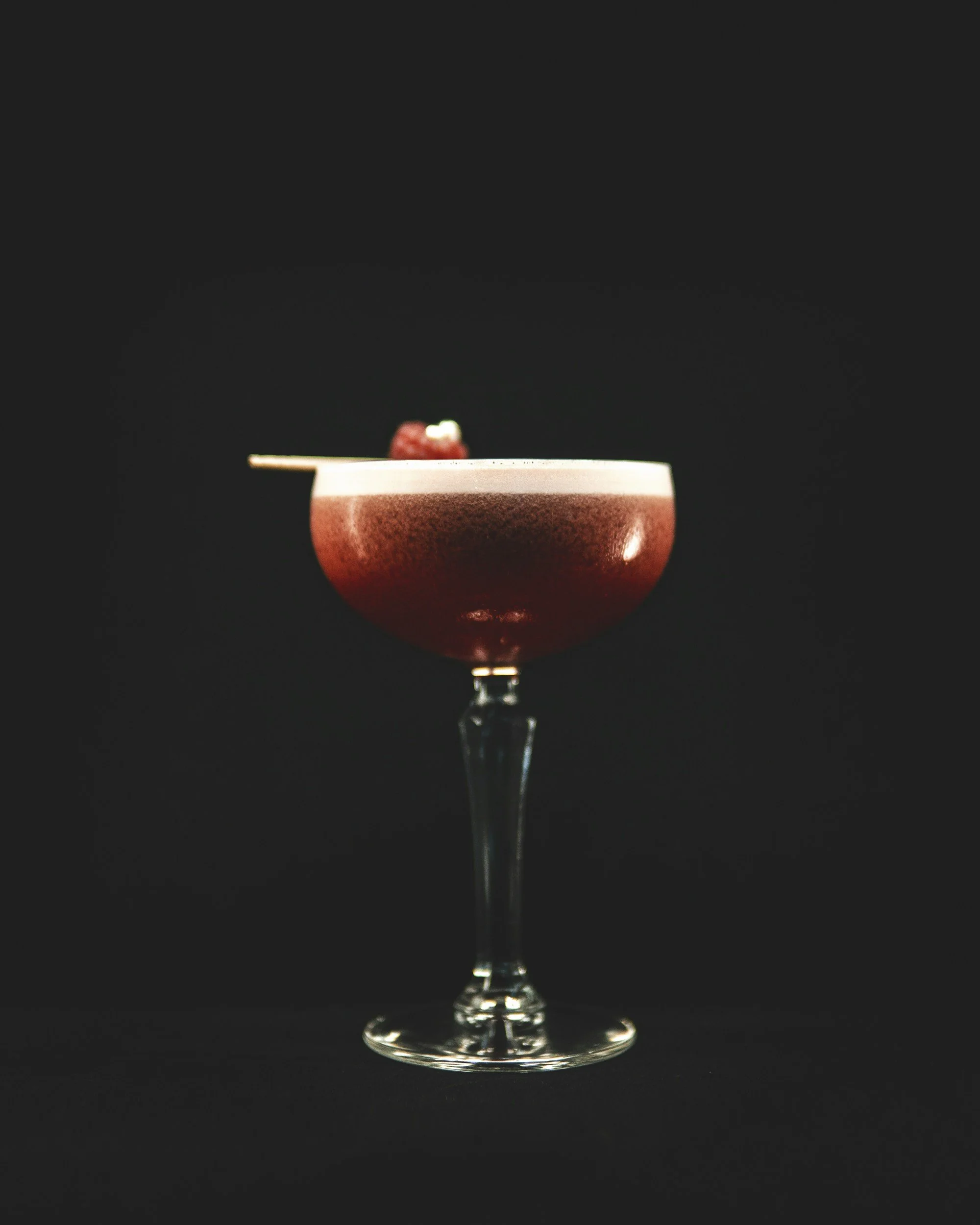A pink cocktail in a coupe glass garnished with a raspberry and a small white edible decoration, against a black background.