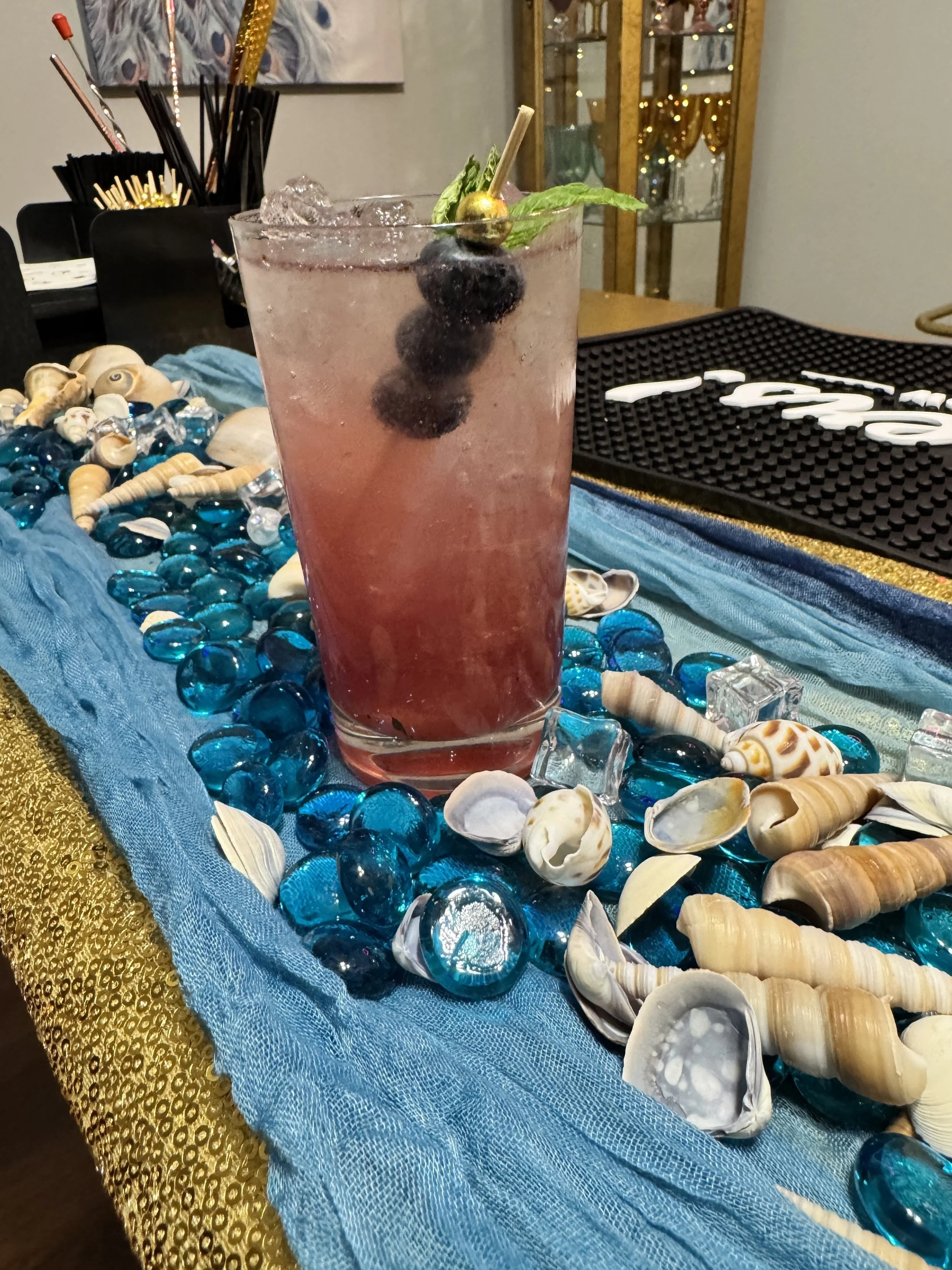 A pink cocktail garnished with blueberries and a sprig of mint, placed on a table decorated with blue and gold fabric, shells, and glass stones, with a bar in the background. offered at MSE Luxe - Atlanta luxury mobile bar service.