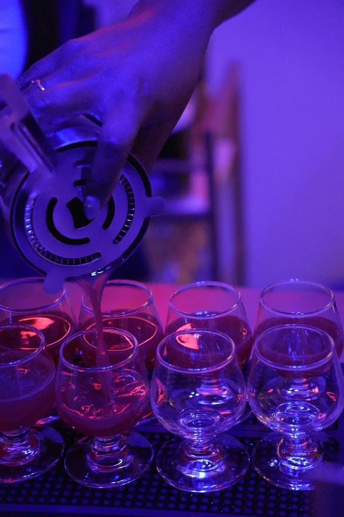 Person pouring pink cocktail into glasses in a dimly lit setting with purple lighting.