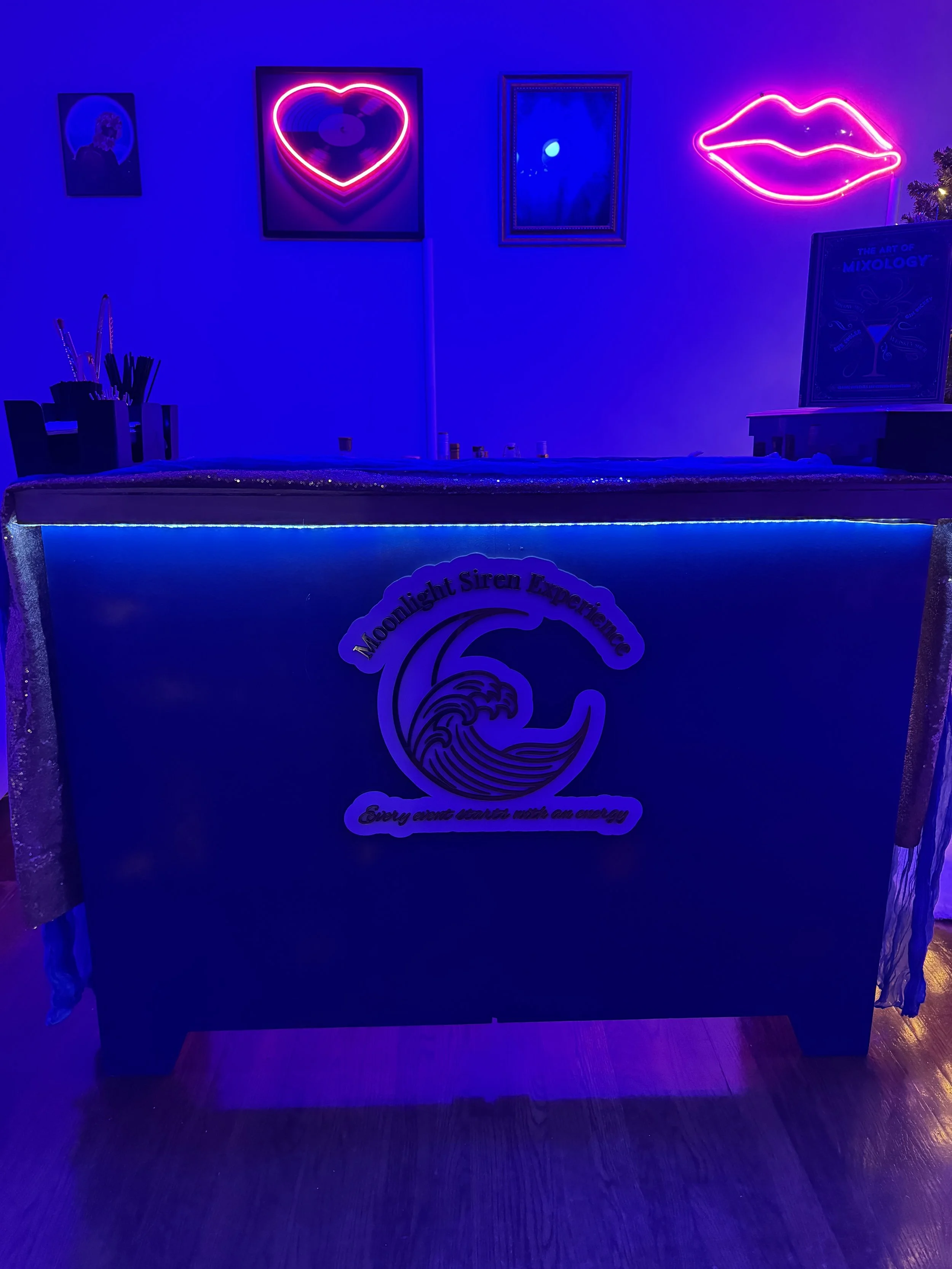 A bar or event table decorated with neon signs of a heart, lips, and art piece on the wall, illuminated mostly in blue and pink neon lights, with a sign reading 'Moonlight Siren Experience' and a wave logo.