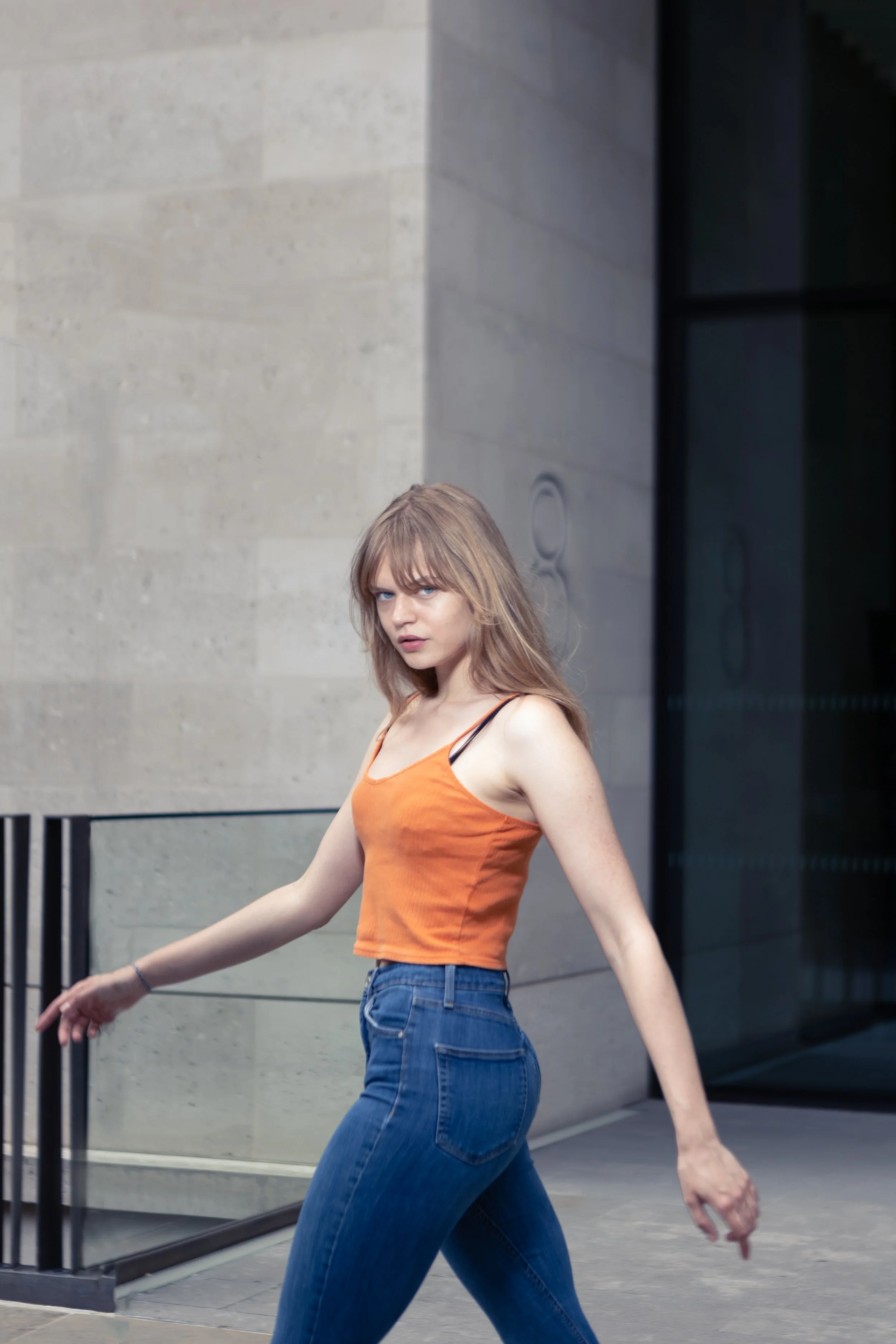An editorial shot of a Young woman with shoulder-length blonde hair and bangs walking outdoors in front of a beige building wall, wearing an orange tank top and blue jeans.