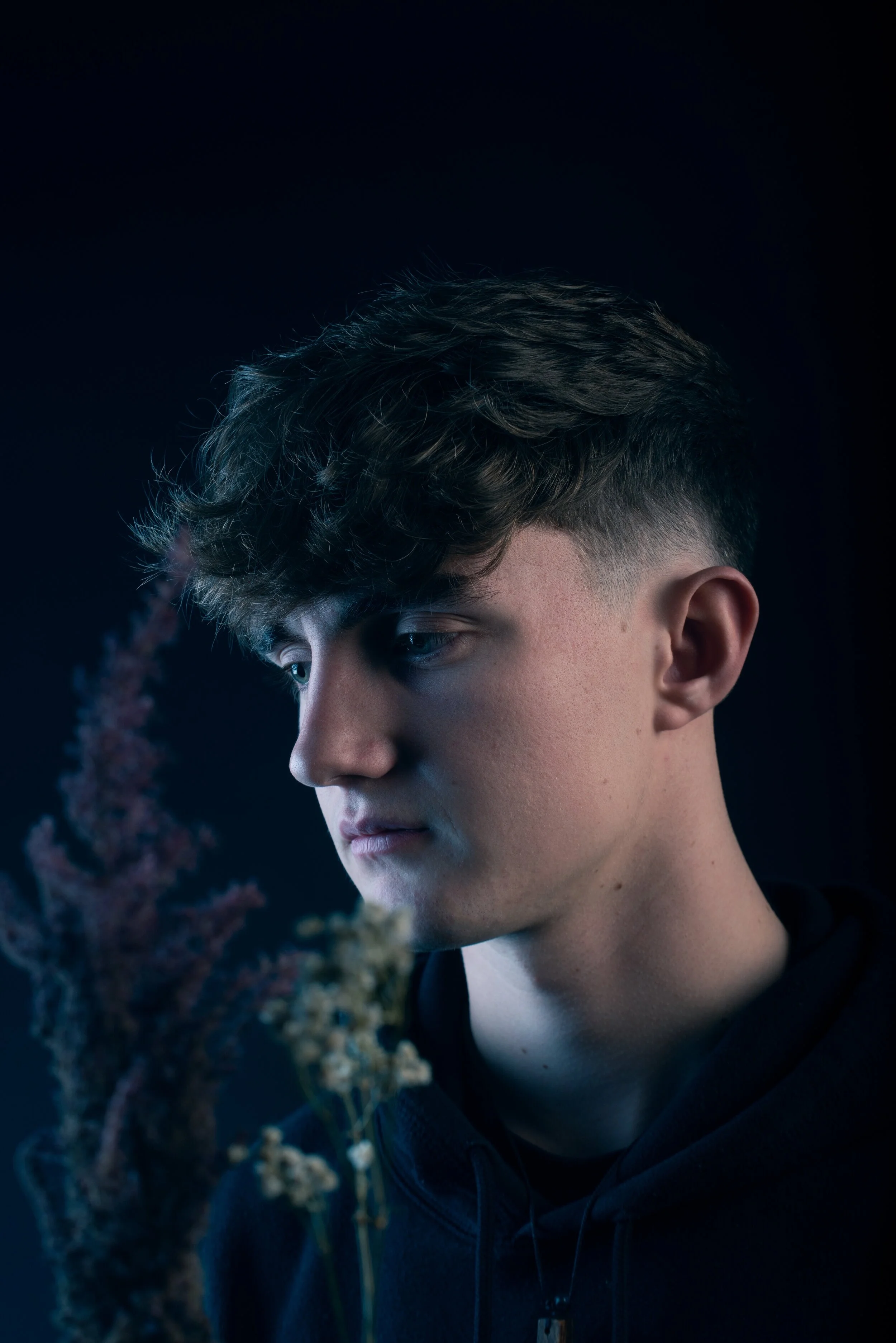 A young man with short, curly brown hair and fair skin gazes downward in a dark setting, with dried flowers in the foreground.