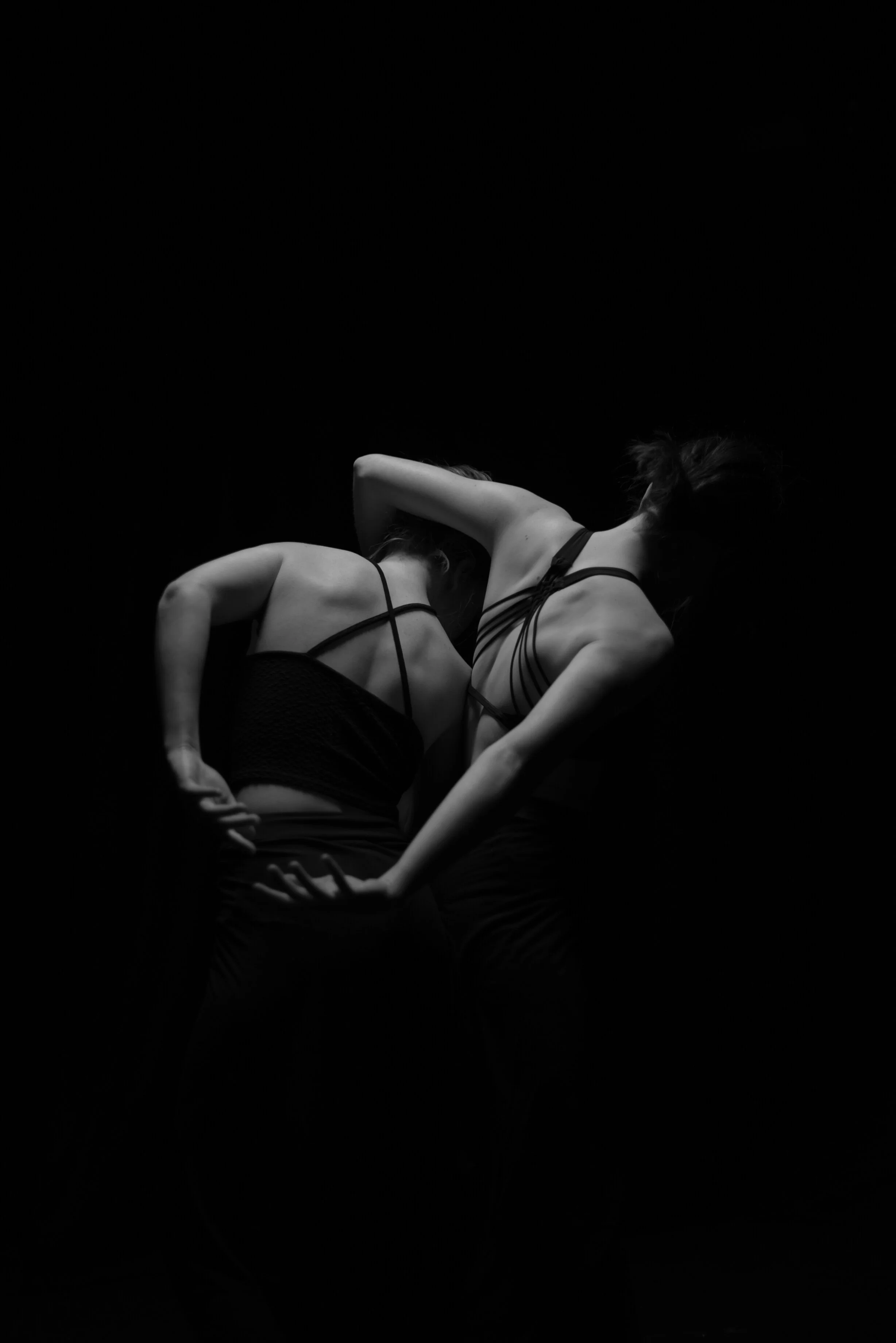 Two women in dark clothing dance or pose against a black background, with dramatic lighting highlighting their backs and arms.