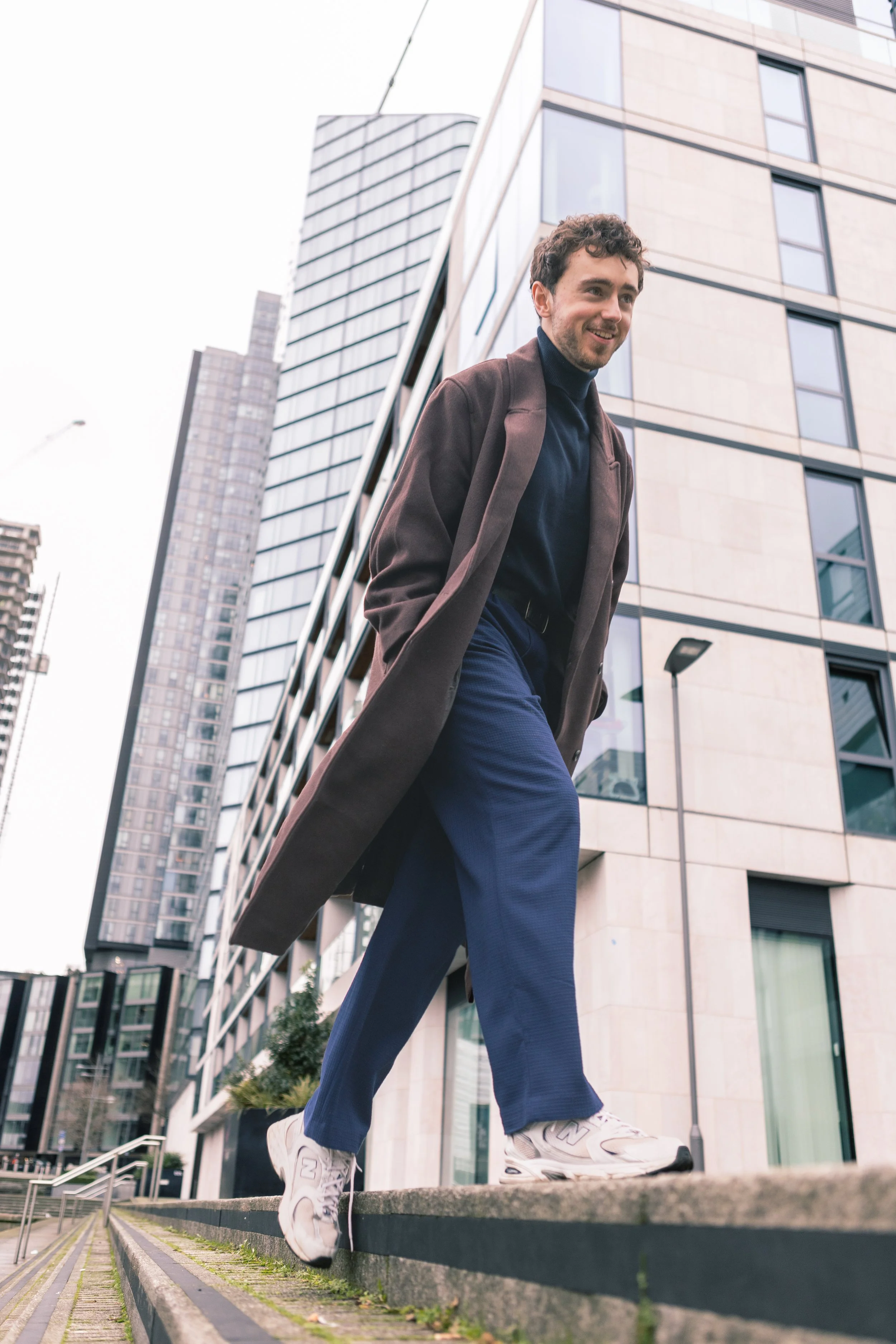 An editorial shoot in the city of a fashionable man in a brown coat, dark shirt, and blue pants walking on a ledge in an urban area with modern buildings in the background.