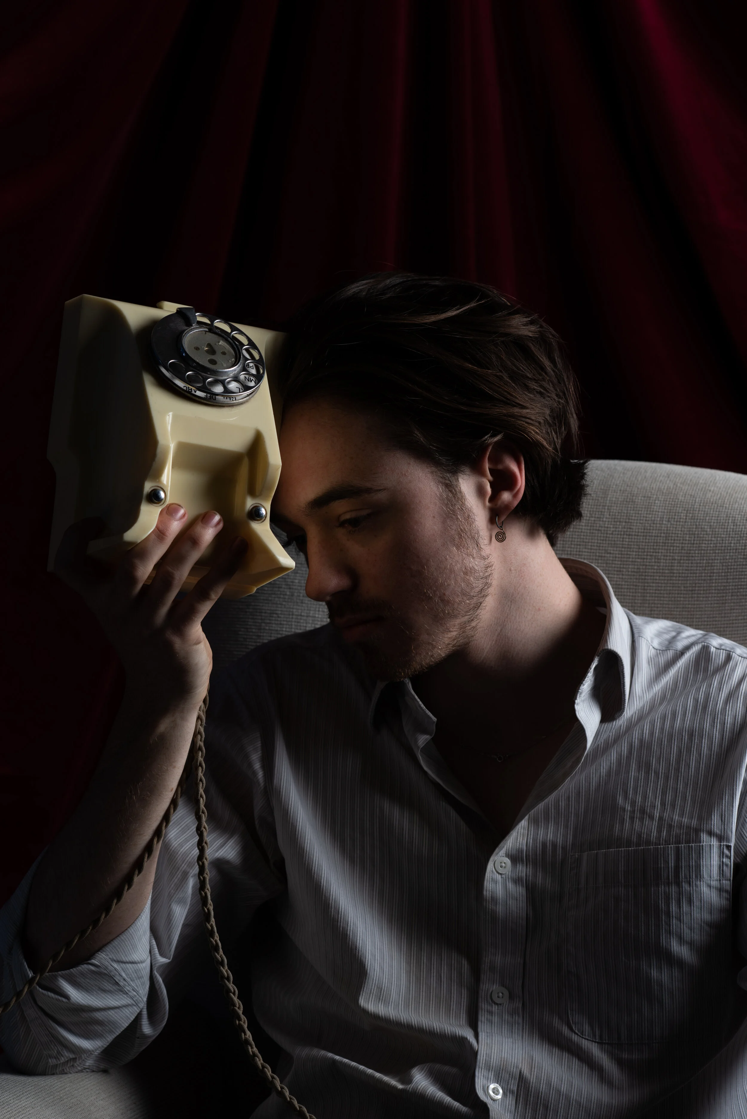 Young man in a striped shirt holding a vintage rotary phone to his head, sitting on a beige chair against a dark red curtain background.