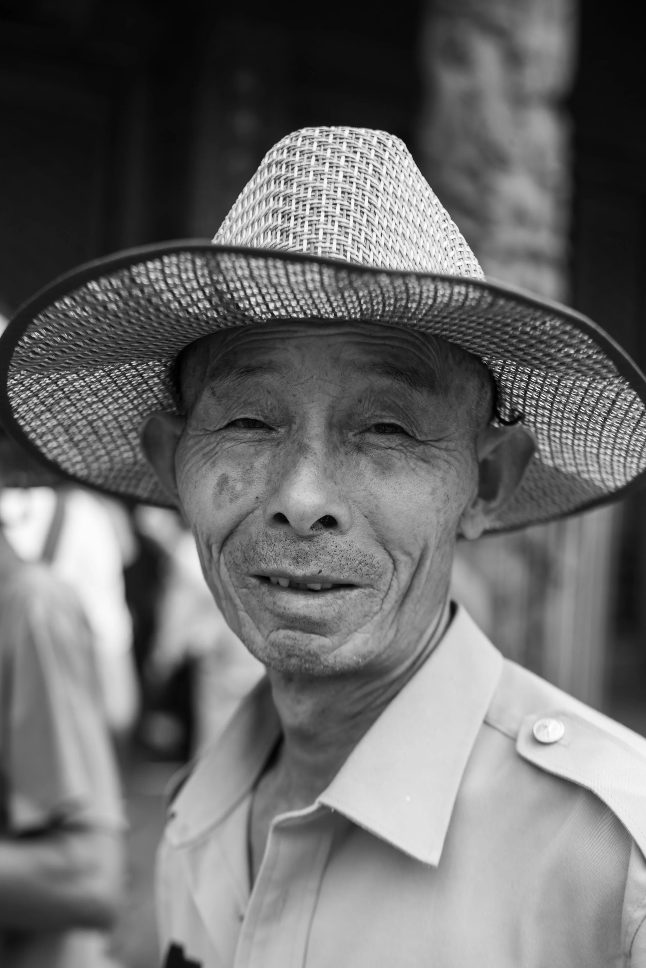 A smiling elderly man wearing a wide-brimmed straw hat and a collared shirt.