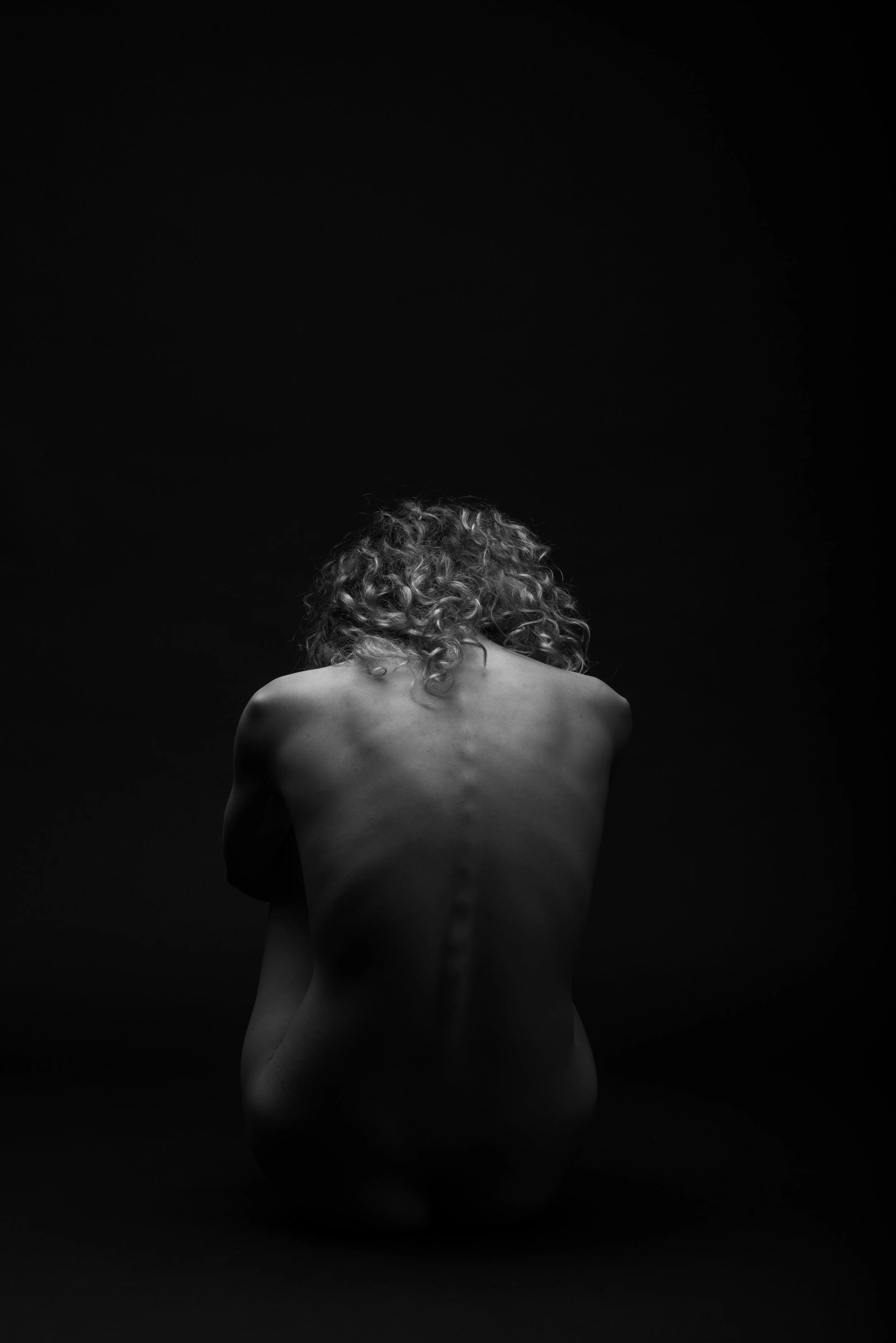 Black-and-white photograph of a woman with curly hair, kneeling with her back to the camera against a dark background.