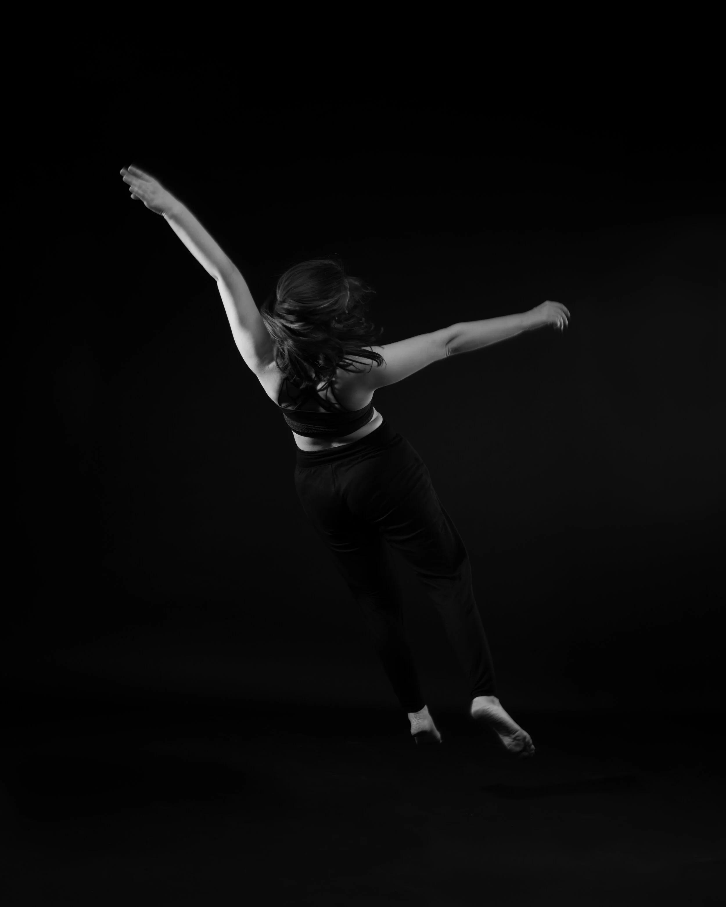 A woman dancing or performing a dance move against a black background, in black athletic clothing.
