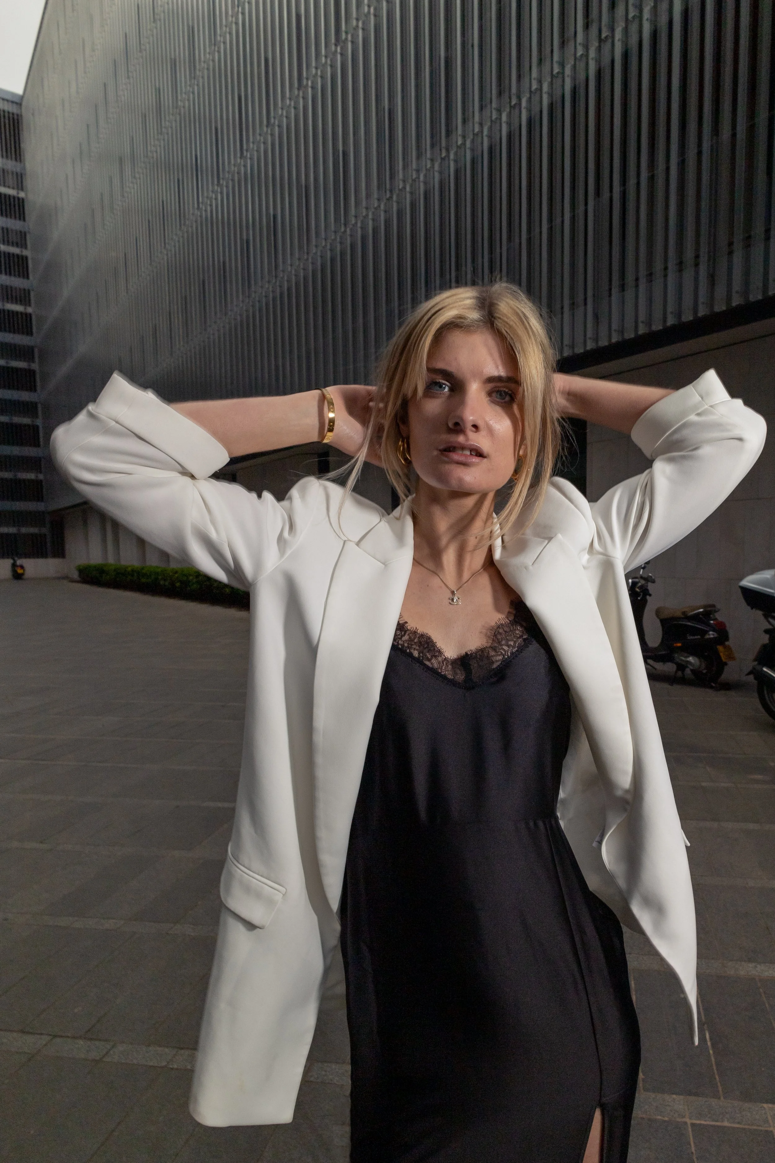 A young woman with blonde hair wearing a black dress with lace detail and a white blazer, standing outdoors at night with her hands behind her head against a modern building.