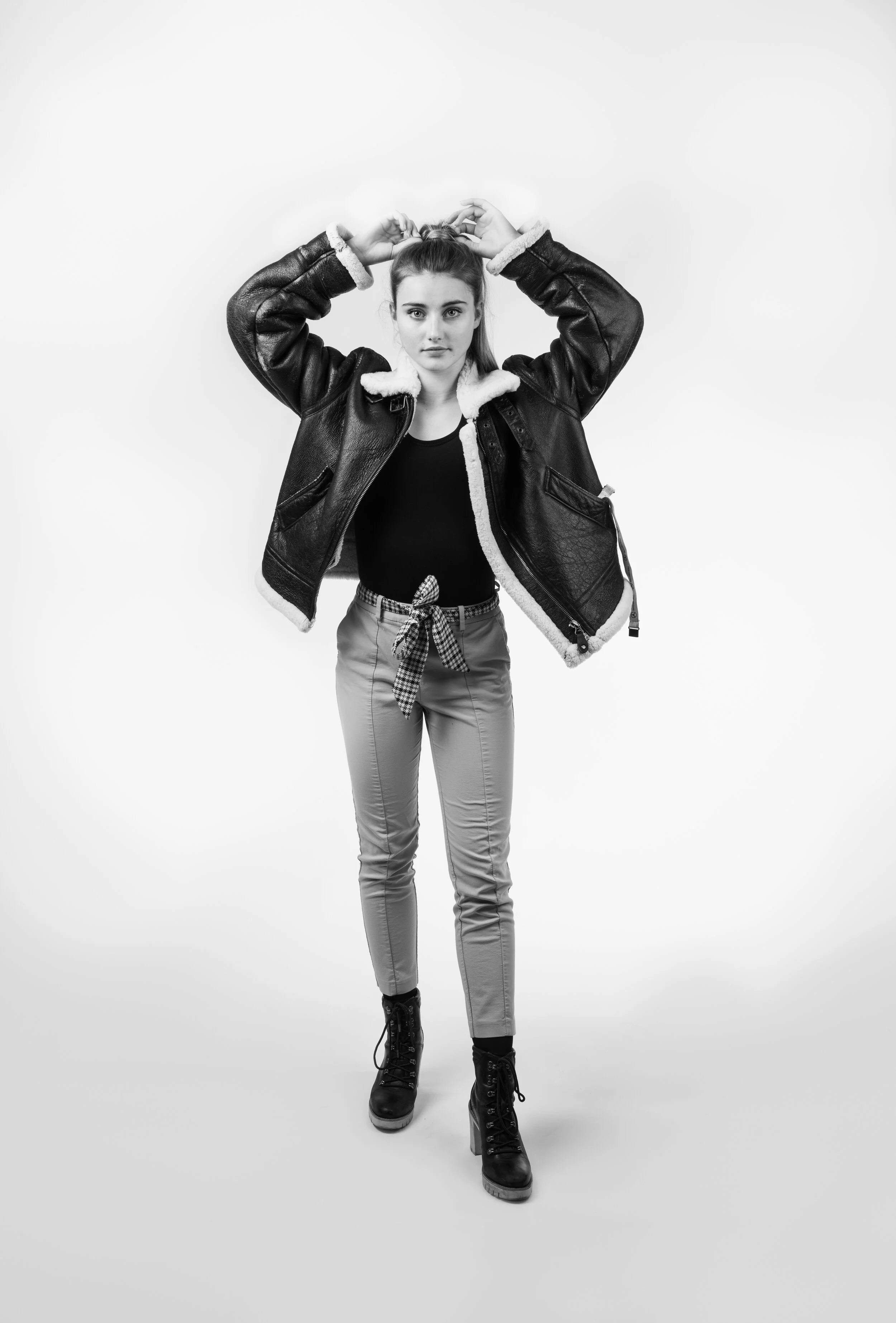 A young woman standing and adjusting her hair in a studio with a plain background, wearing a black shirt, plaid tied belt, light pants, black boots, and a black jacket with shearling lining.