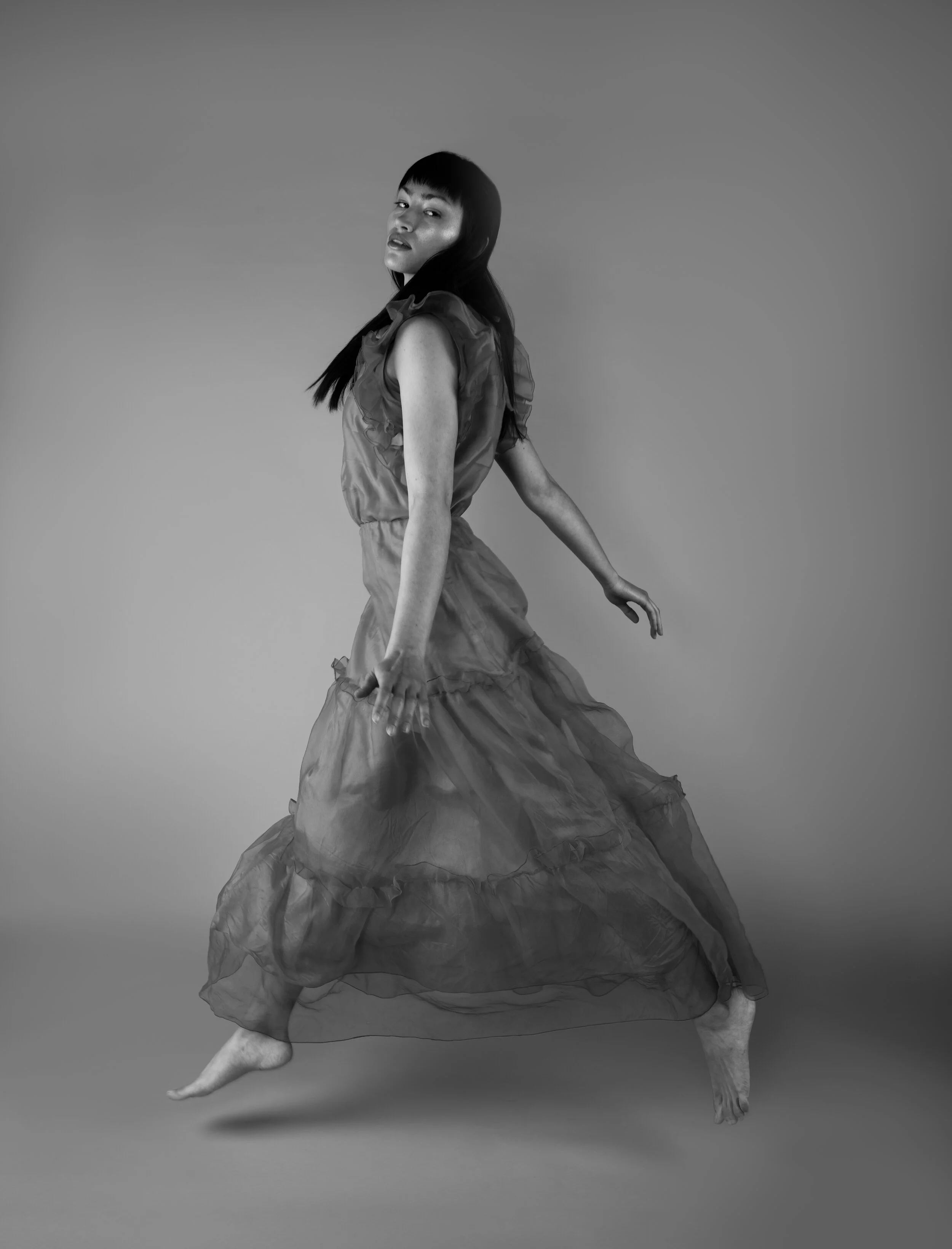 An editorial studio portrait, black-and-white photo of a woman in a flowing dress, captured mid-movement, with a neutral background.