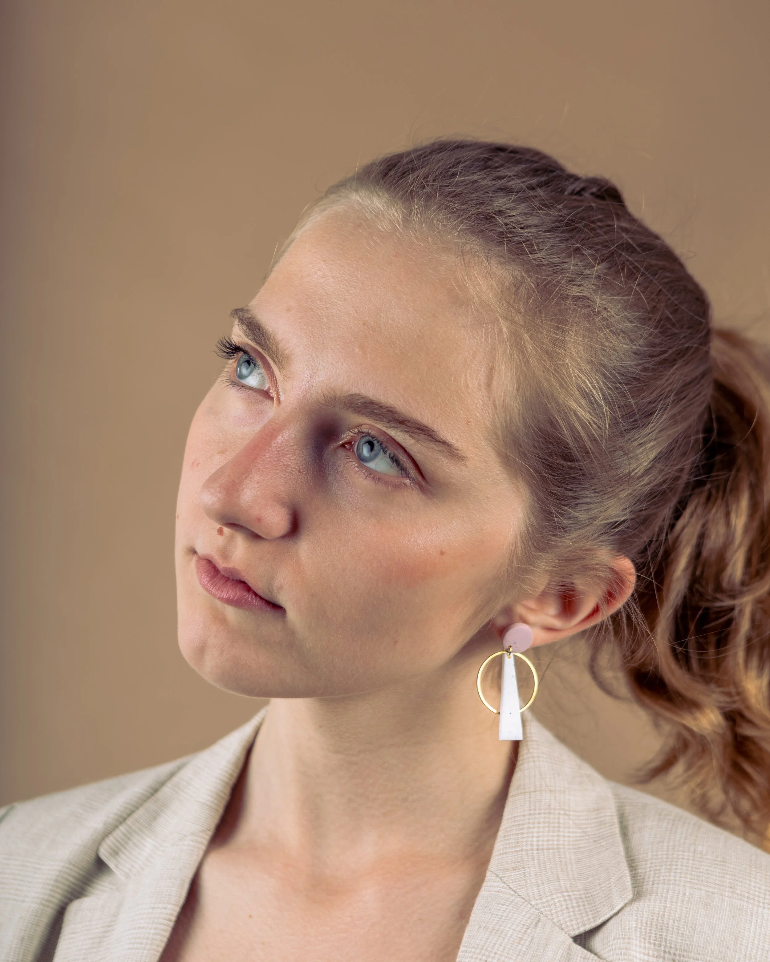 A studio shoot for a jewellery brand with a close-up of a woman with blue eyes and light brown hair in a ponytail, wearing a beige blazer and a large decorative earring, looking thoughtfully upward.