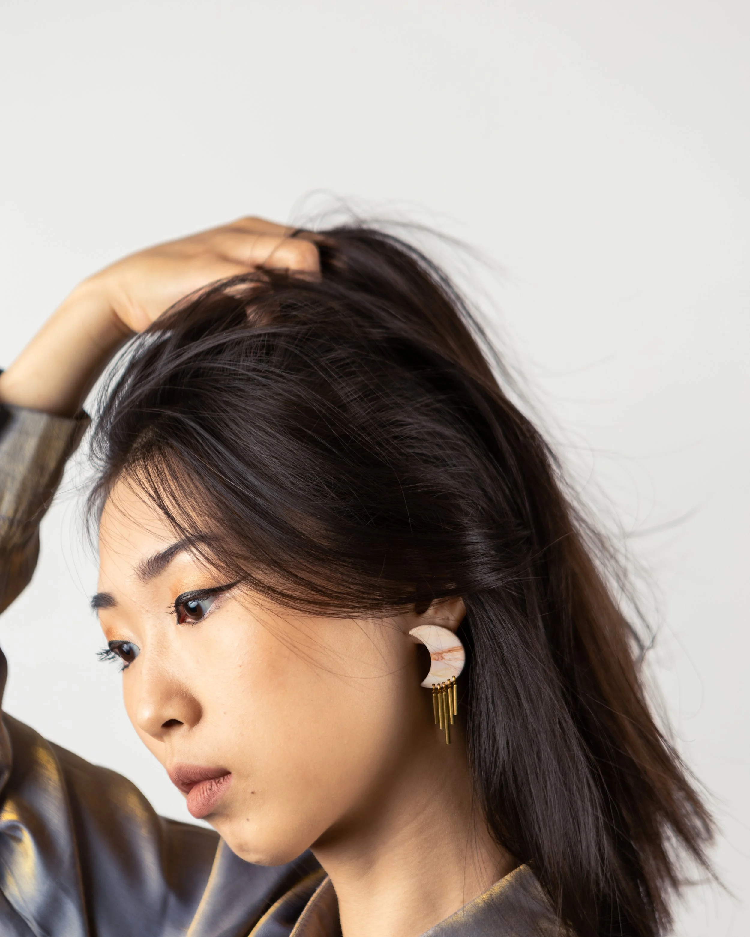 A studio shoot for a jewellery brand with a woman with dark hair, wearing gold earrings, looking down and touching her hair.