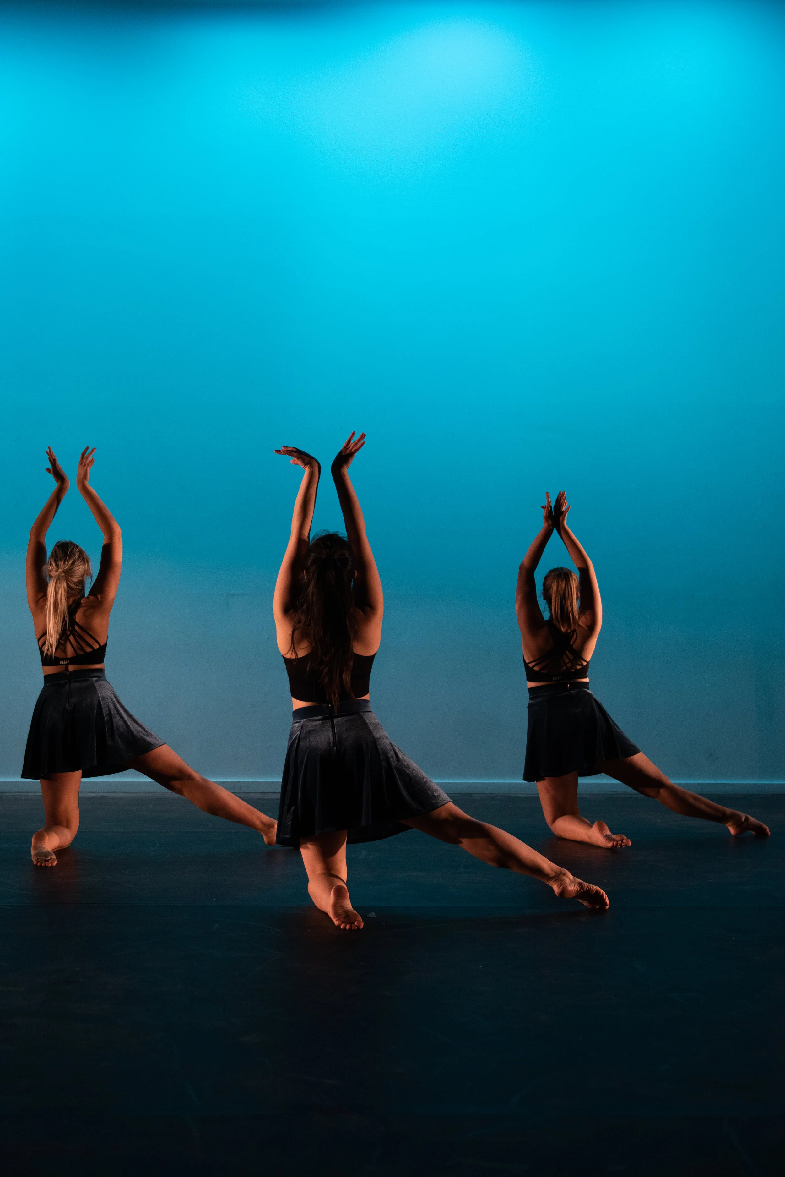 Three women in black dance costumes performing a dance routine on stage, with their arms raised and legs extended, against a bright blue background.