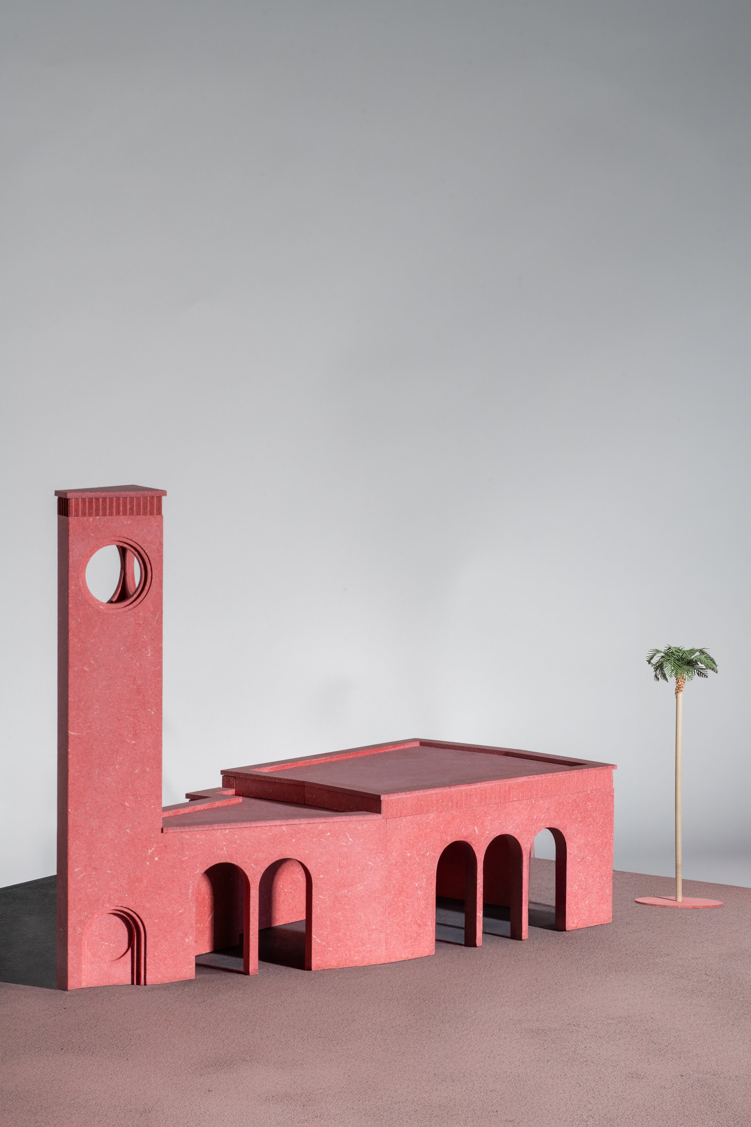 A miniature architectural model of a pink building with a tower, arches, and a flat roof, along with a small artificial palm tree.
