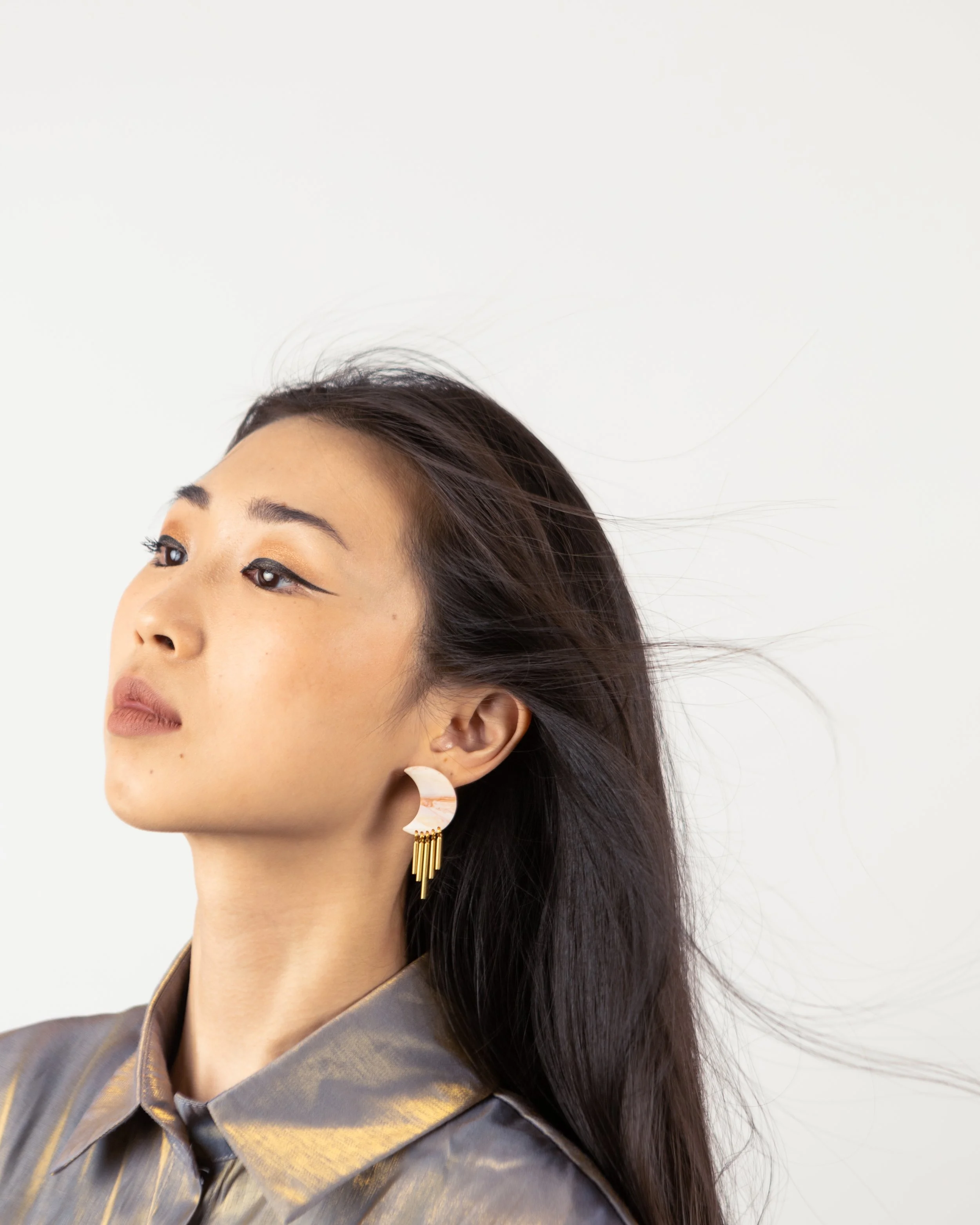 A woman with long dark hair wearing a metallic silver collared shirt and beige and pink geometric earrings.