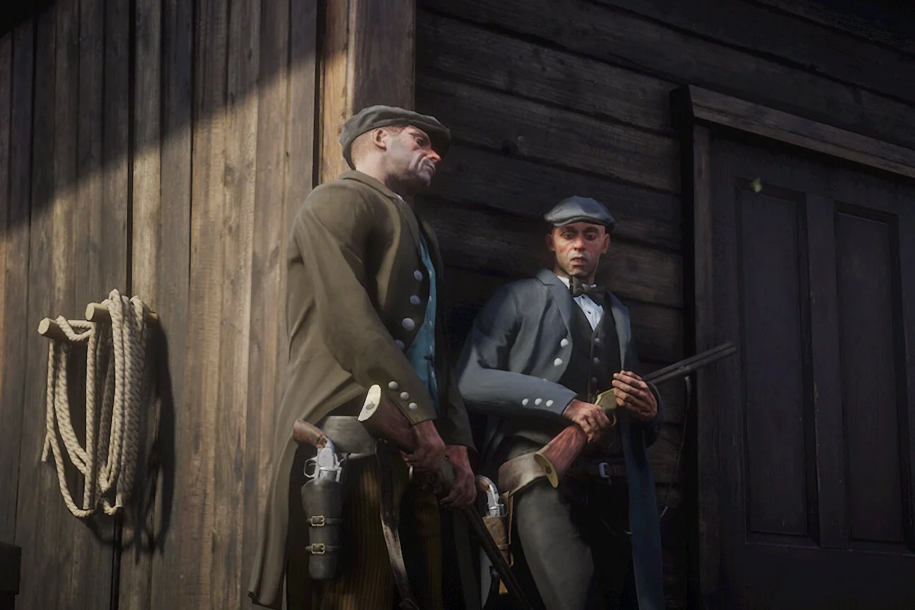 Two men dressed as soldiers in historical military uniforms, standing inside a wooden building with dark walls, one holding a rifle and the other holding a pistol.